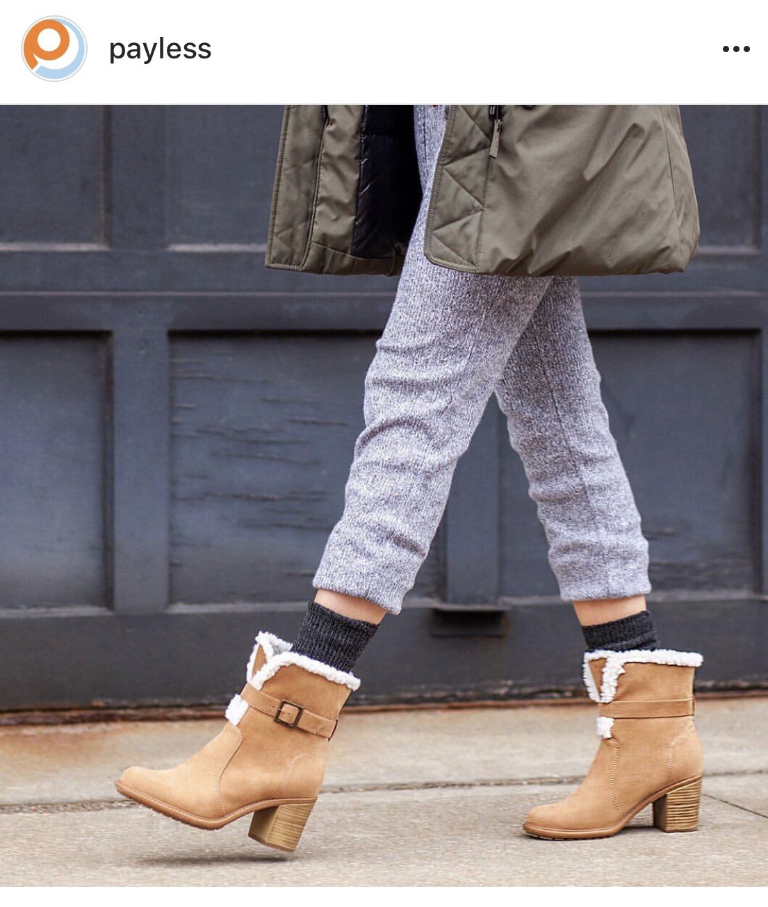 Close-up of a person's legs wearing gray textured pants, black socks, and tan shearling-lined ankle boots with a buckle, walking on concrete sidewalk with a dark gray garage door in the background.