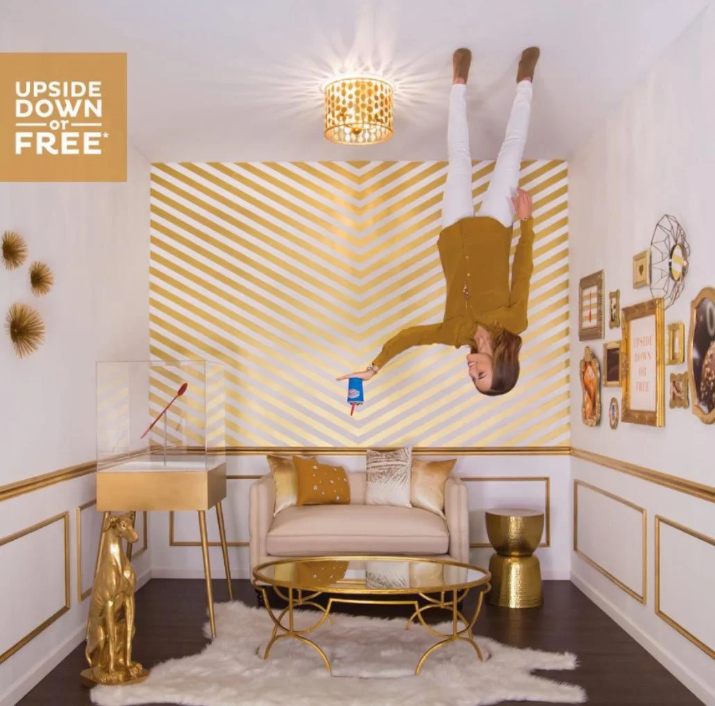 A woman is upside down in a room with gold and white decor. She is holding a cup with a straw and a small blue container. The room features a gold deer statue, a glass display case, a gold side table, a cream sofa with pillows, gold framed art on the