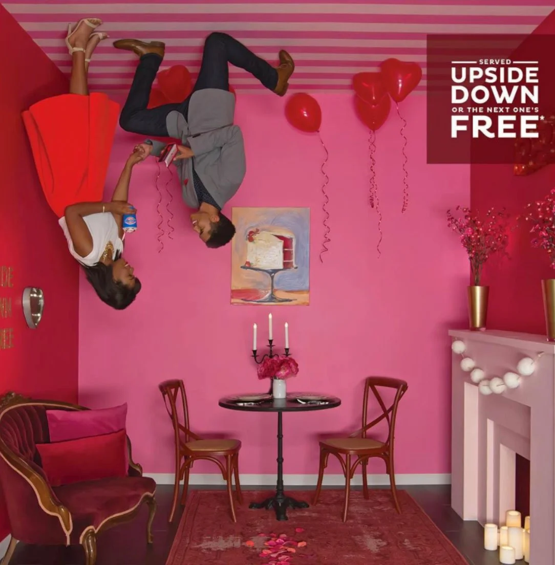 Room decorated in pink with balloons and candles, featuring a woman and a man lying upside down on a pink couch, with a sign saying 'Served upside down or the next one's free'.