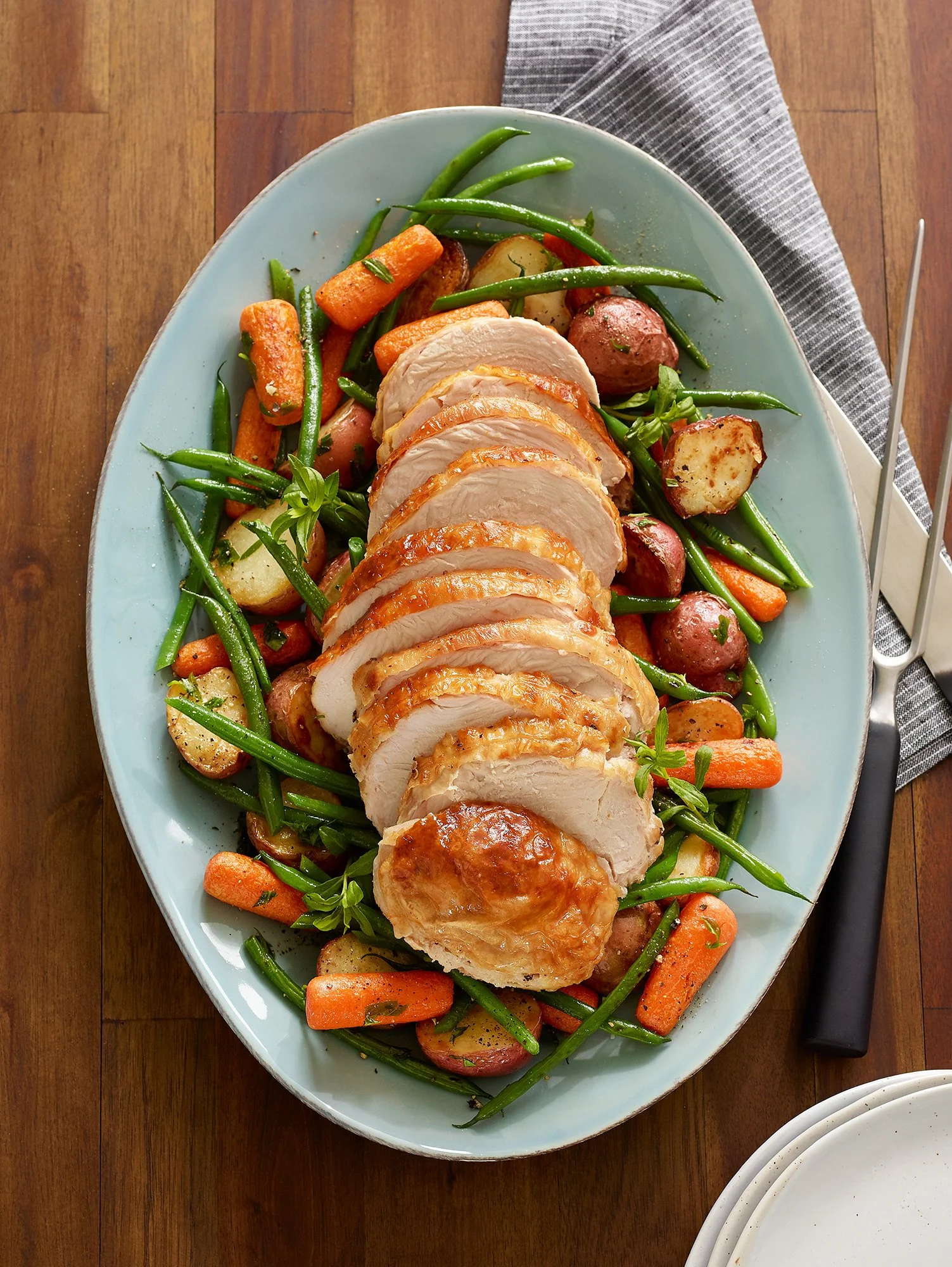 A plate of sliced roasted chicken breast on a bed of green beans, carrots, potatoes, and small red potatoes on a wooden table.