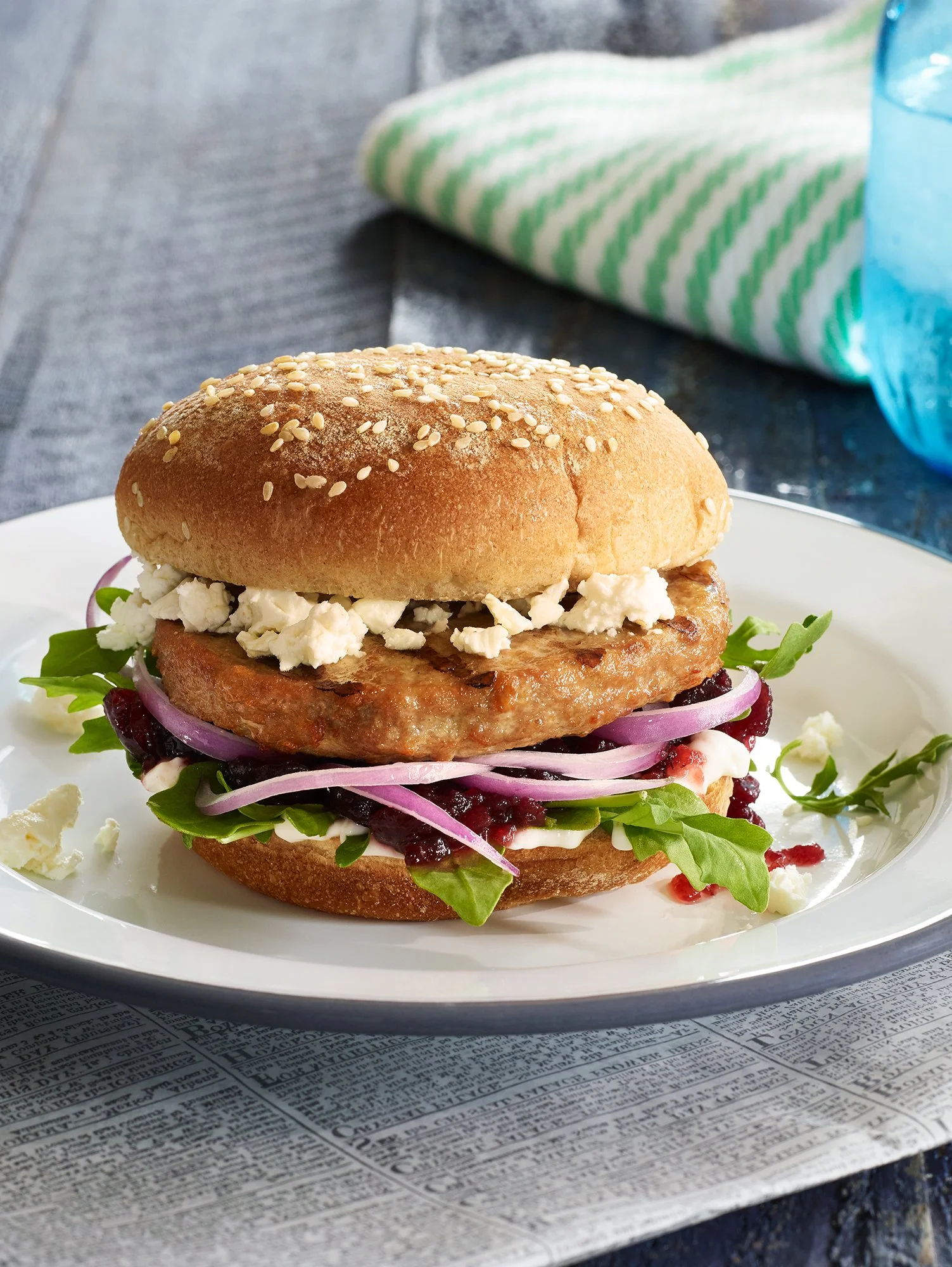 A sandwich on a white plate with leafy greens, purple onions, red berries, feta cheese, and a toasted bun, resting on a sheet of newspaper.