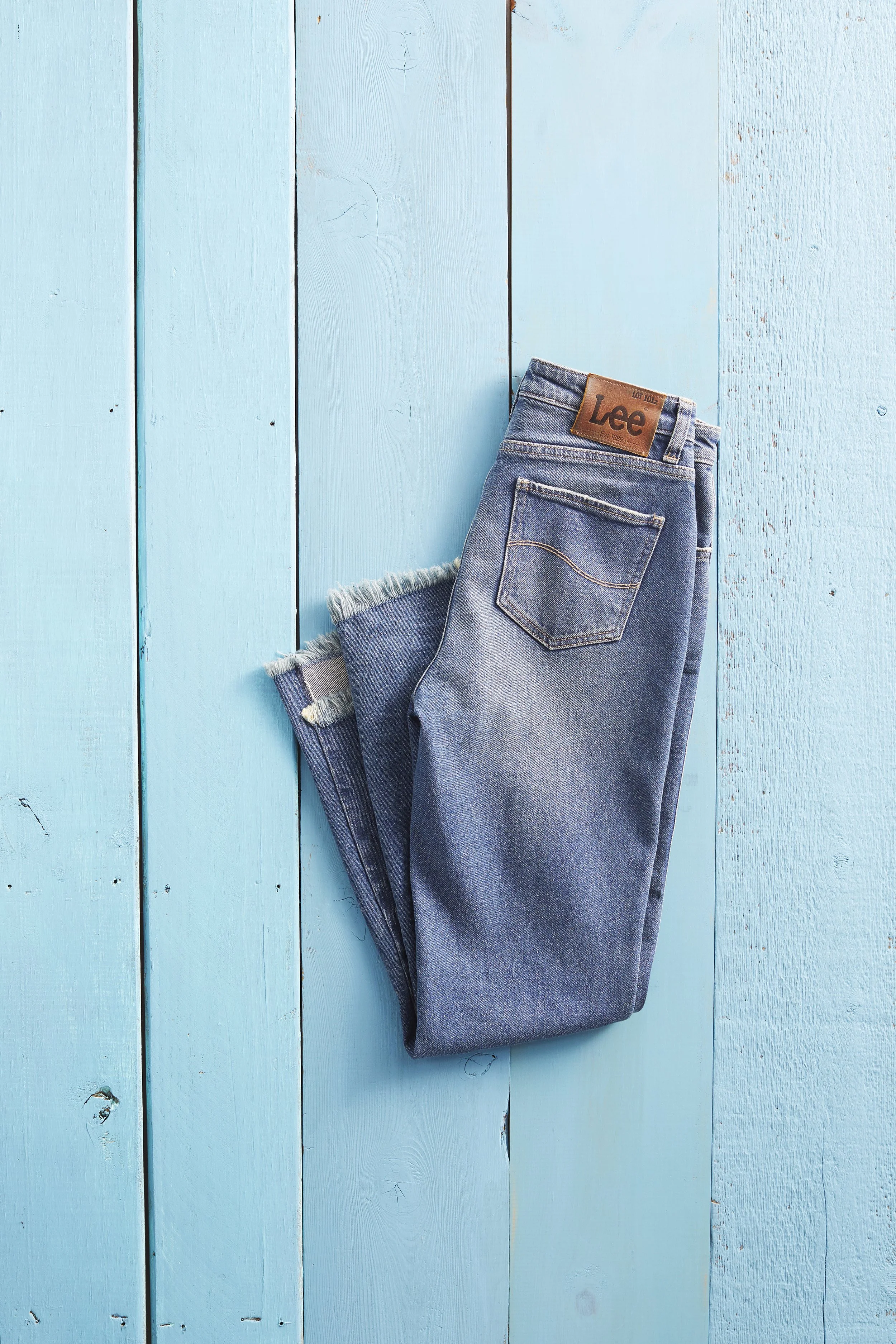 A pair of folded blue denim jeans with a brown leather Lee brand patch on the waistband, resting on a light blue wooden surface.