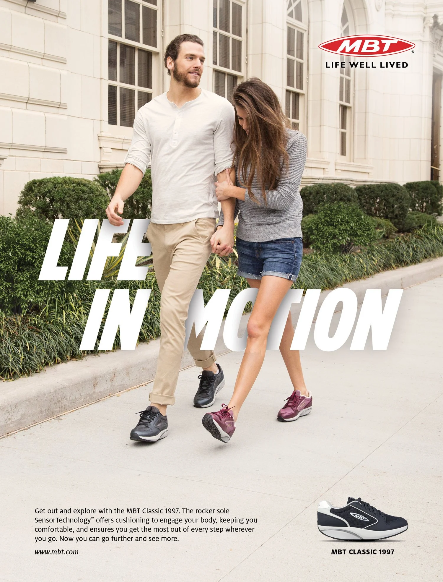 A man and woman walking on sidewalk holding hands near a large white building with windows and greenery, with an advertisement for MBT shoes featuring a pair of black shoes and promoting comfort and cushioning technology.