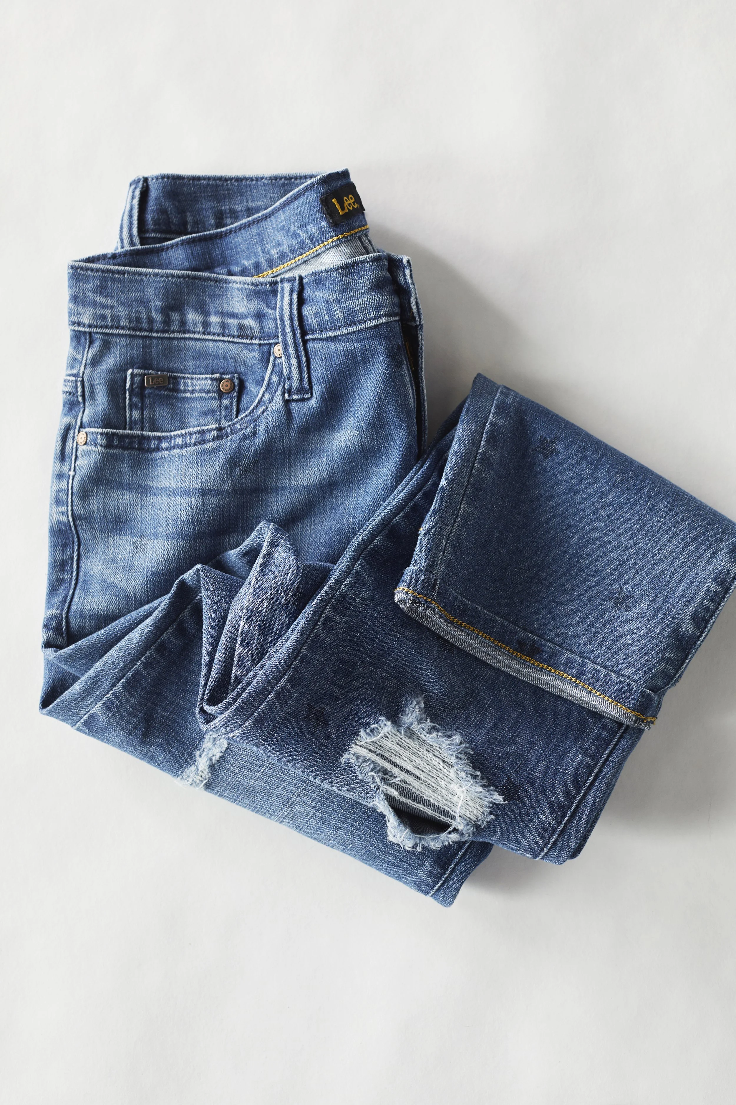 A pair of blue denim jeans with a frayed hole in the knee, folded on a plain white surface.
