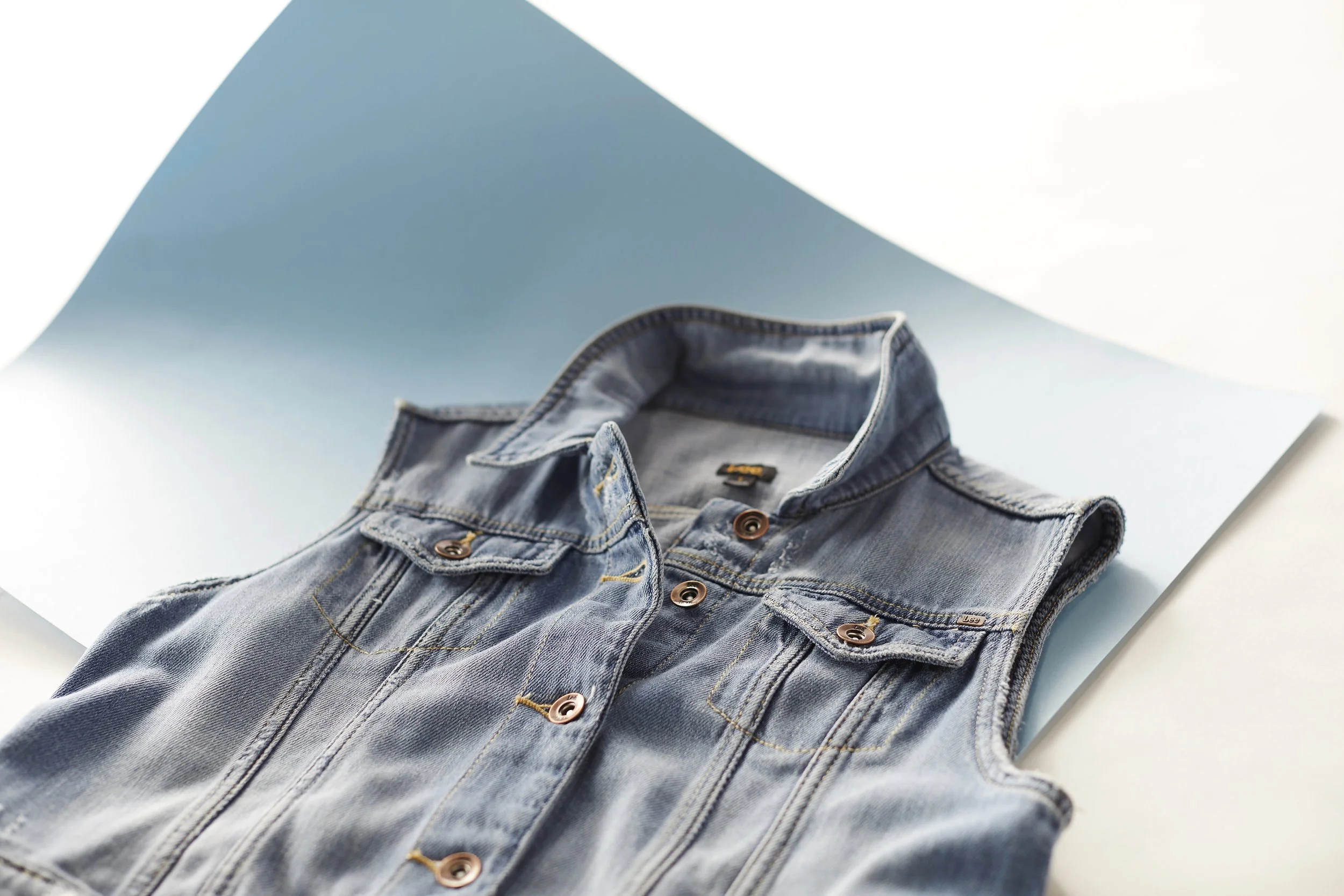 Close-up of a folded blue denim jacket lying on a white surface with a light blue backdrop.