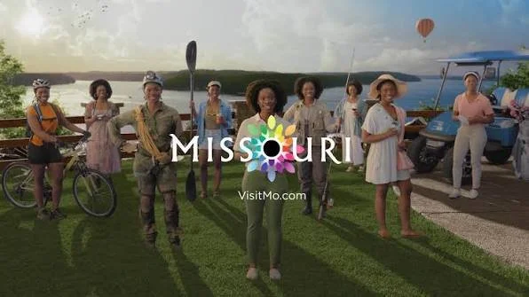 Group of diverse women standing on grassy area with water and a hot air balloon in background, some holding a paddle, with 'MISSOURI' text and VisitMo.com logo overlayed.