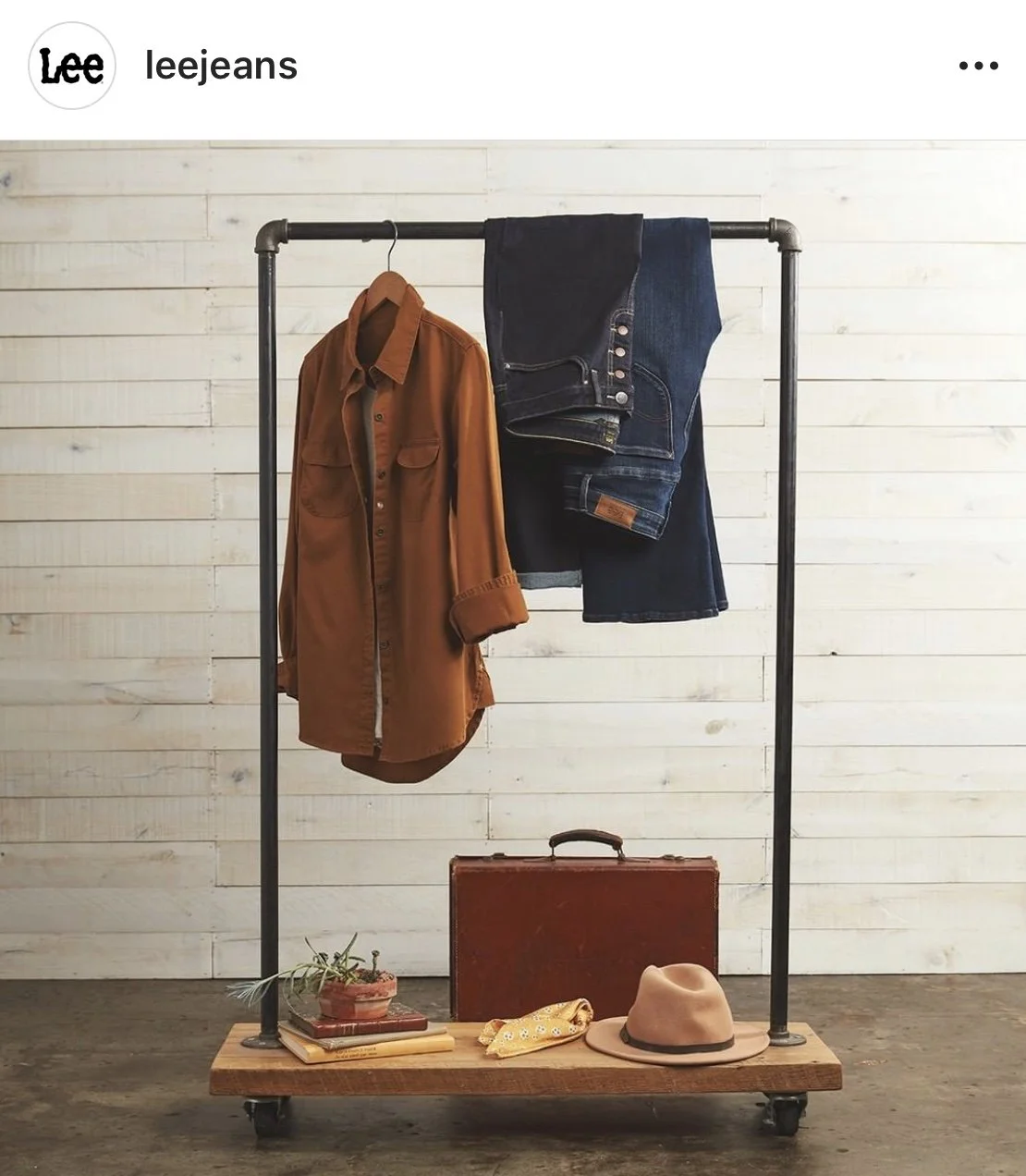 Clothing rack with a brown shirt, a pair of blue jeans, a suitcase, a hat, a plant, a yellow bandana, and books against a white wooden wall.