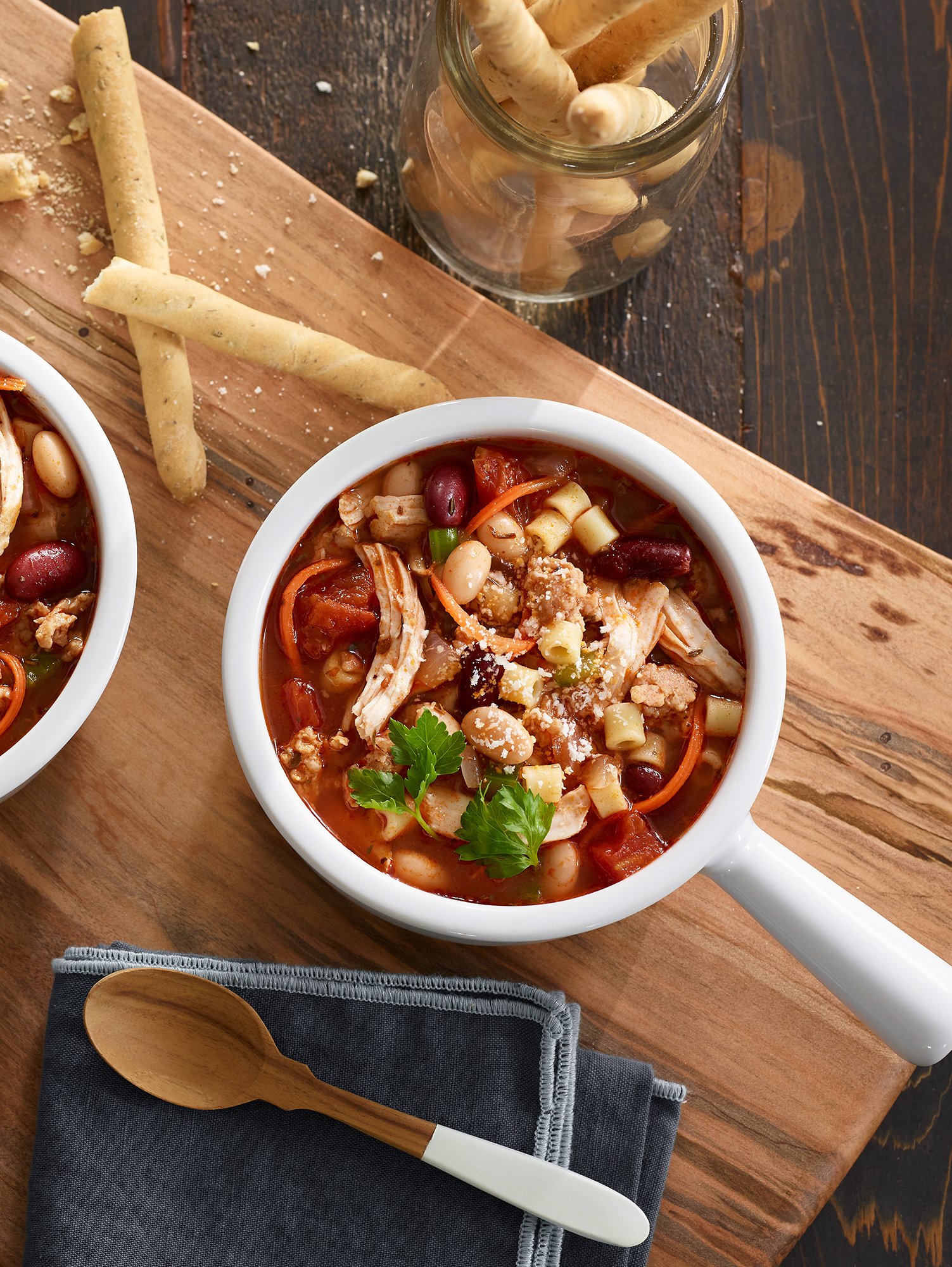 Bowl of chicken tortilla soup garnished with cilantro, served on a wooden table with breadsticks in a glass jar, a wooden spoon, and a navy napkin with a white-handled spoon.