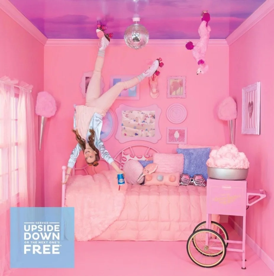The room is decorated in shades of pink and purple with a pink bed, pink fluffy pillows, and pink wall art, featuring a girl hanging upside down from the ceiling with a drink in her hand, and a pink poodle plush toy on a pink cart next to a pink bed, a disco ball hanging from the ceiling, and a pink snow cone lamp on either side of the bed.