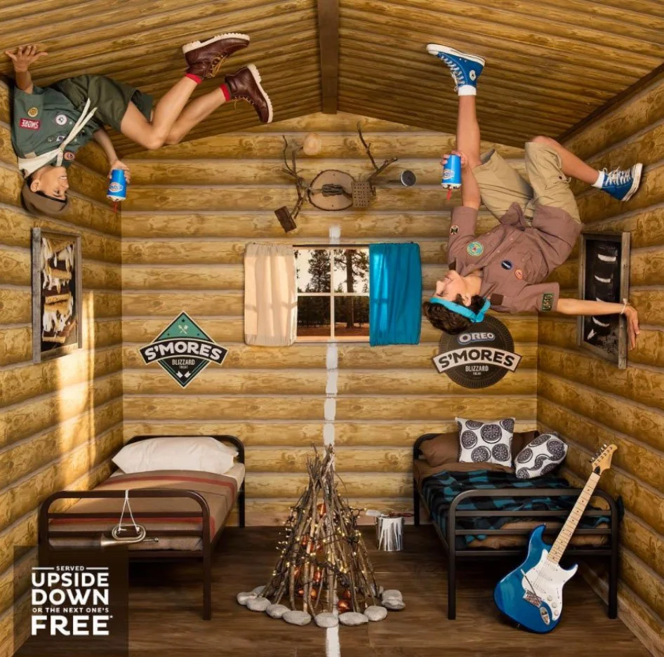 Inside a log cabin with two beds, one with a plaid blanket and the other with patterned pillows, a faux campfire made of sticks and rocks, a window with blue and white curtains, and signs that say 'S'mores Blizzard' and 'S'mores Oreo Blizzard.' There