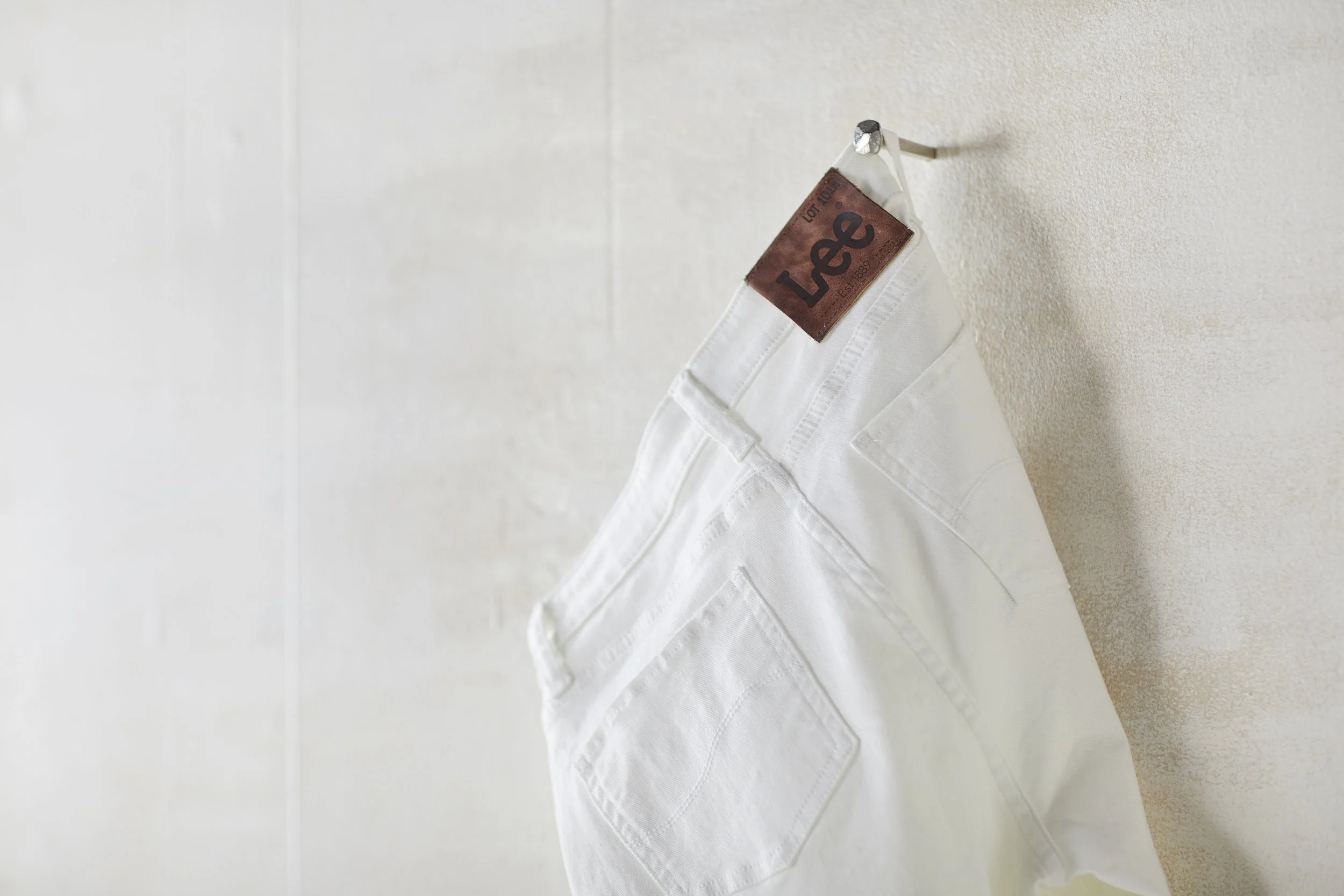 White pair of jeans hanging on a wall hook with a leather patch labeled 'Levi' near the waistband.