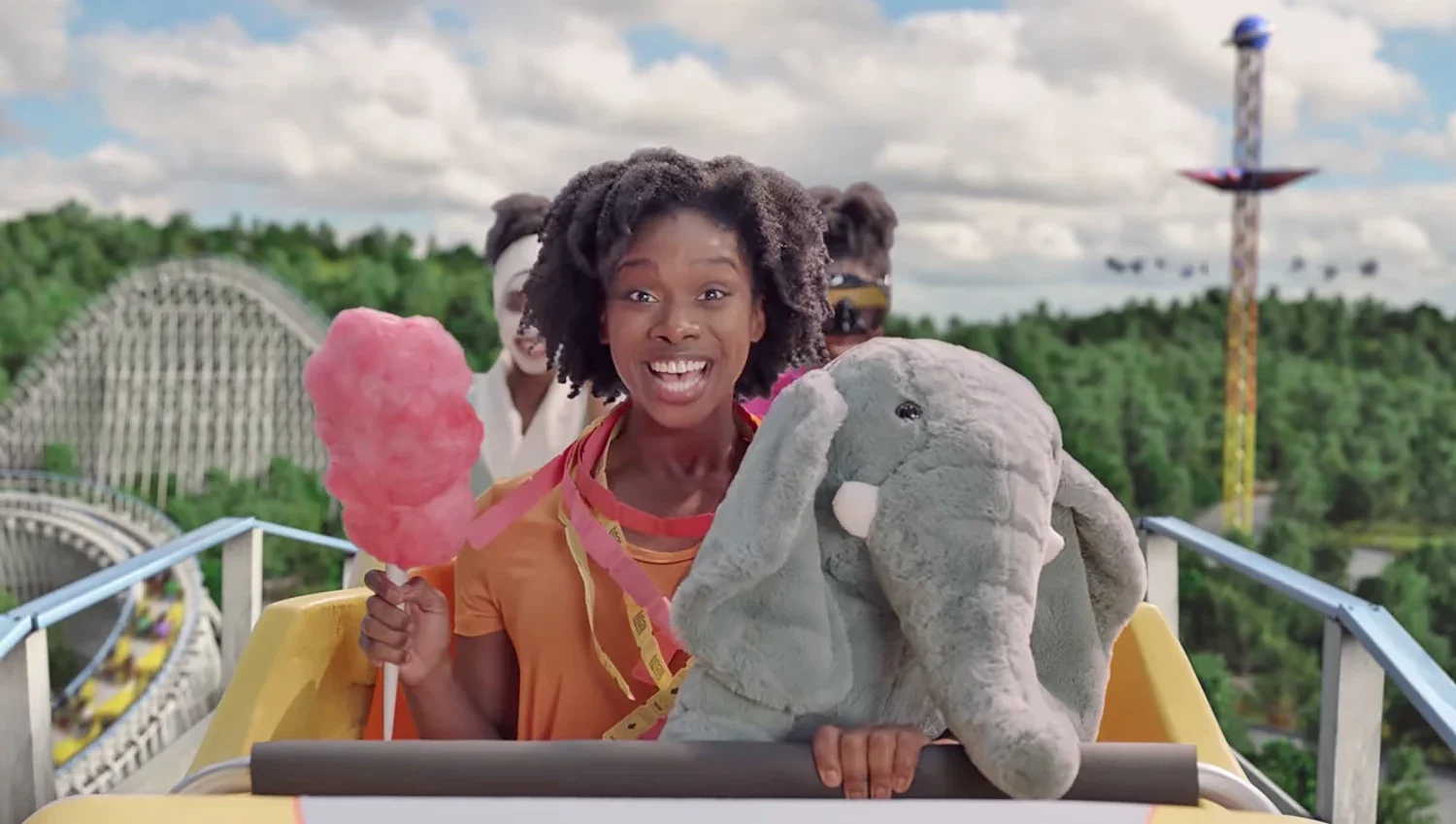 A woman with curly hair smiling while riding a roller coaster at an amusement park, holding a pink cotton candy and a plush elephant. Other riders are visible behind her, with roller coaster tracks and amusement park rides in the background.