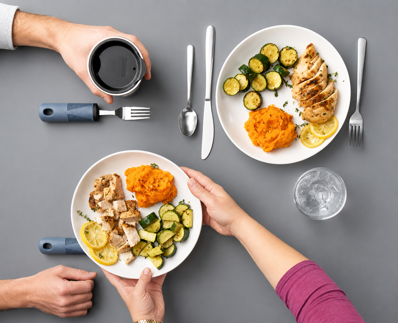 People sharing a meal with plates containing sliced chicken, zucchini, mashed sweet potatoes, and lemon slices, along with a glass of water, on a gray table.