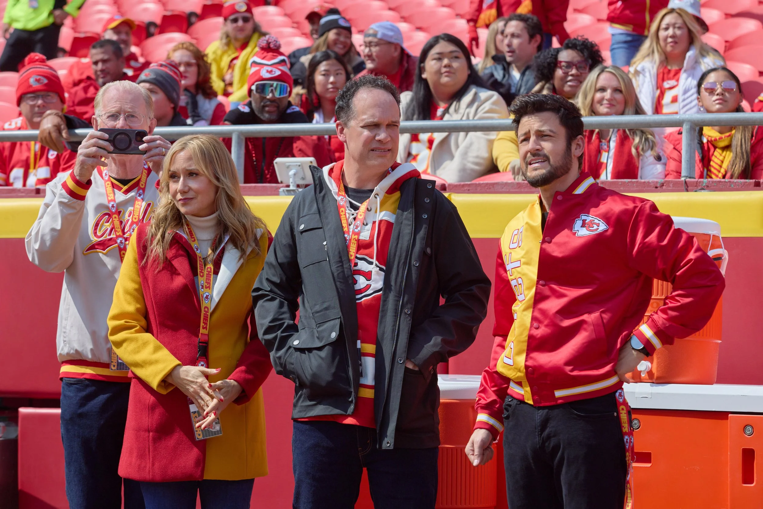 A group of diverse sports fans, some wearing Kansas City Chiefs team apparel, are seated in stadium seats, with three people standing in front. Some are taking photos or looking around, with a red and yellow stadium setting.