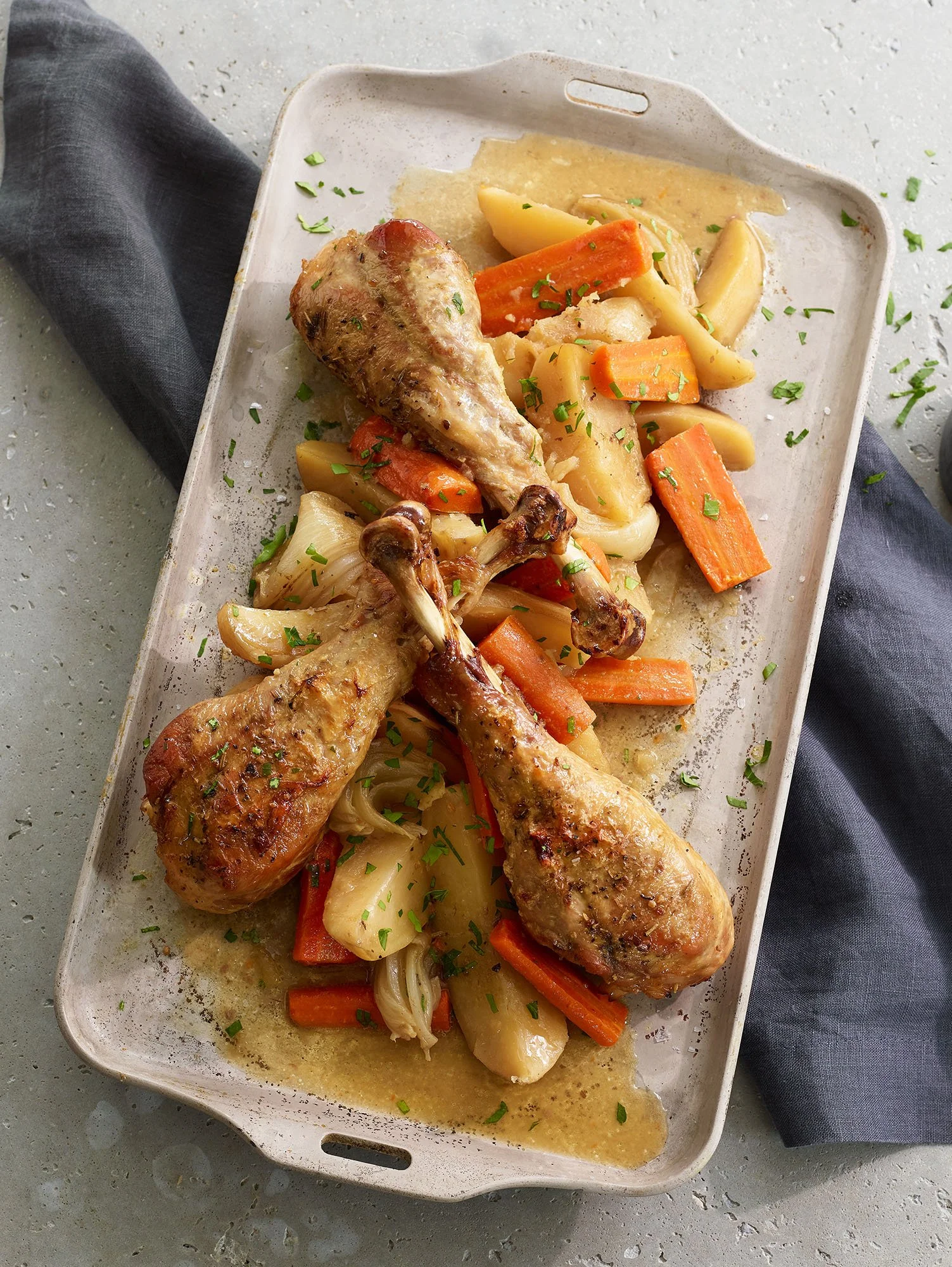 A white rectangular dish containing cooked chicken drumsticks with roasted vegetables, including carrots, onions, and potatoes, garnished with chopped green herbs, and a light gravy.