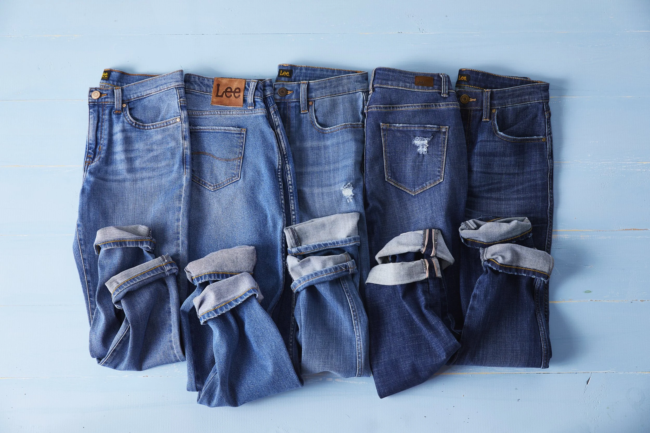 Five pairs of folded blue jeans with different shades and styles laid out on a light blue wooden surface.