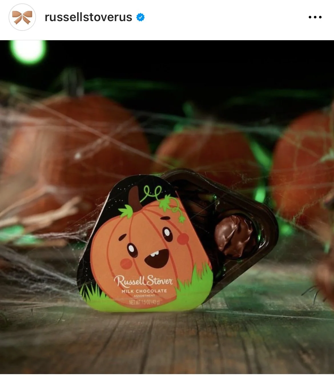 A chocolate candy in a Halloween-themed wrapper featuring a cute cartoon pumpkin with a smiling face and green vine decorations. The wrapper has the brand name Russell Stover and is placed on a wooden surface with spider webs, and blurred orange pump