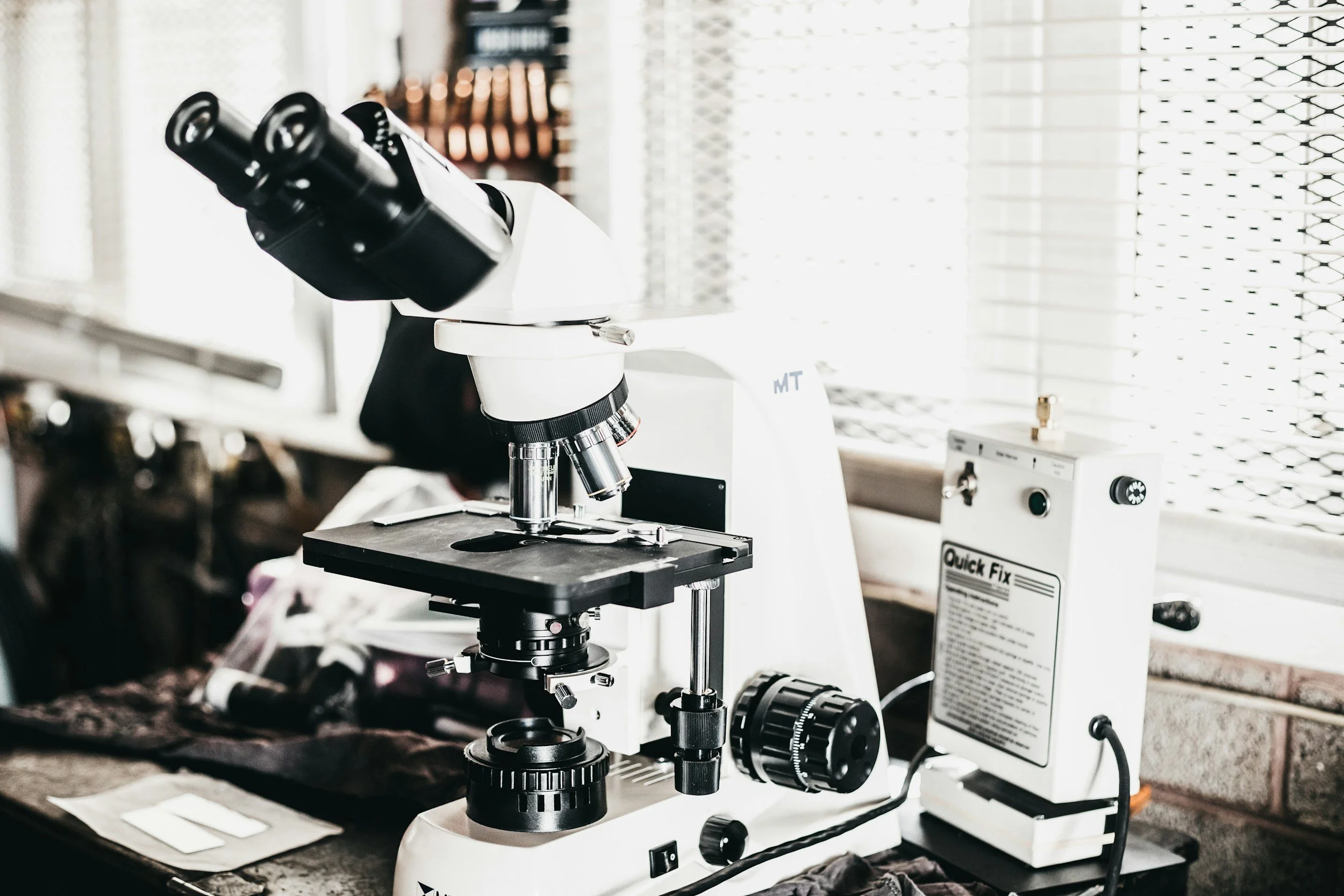A white microscope with black and metallic components placed on a countertop near a window with blinds.