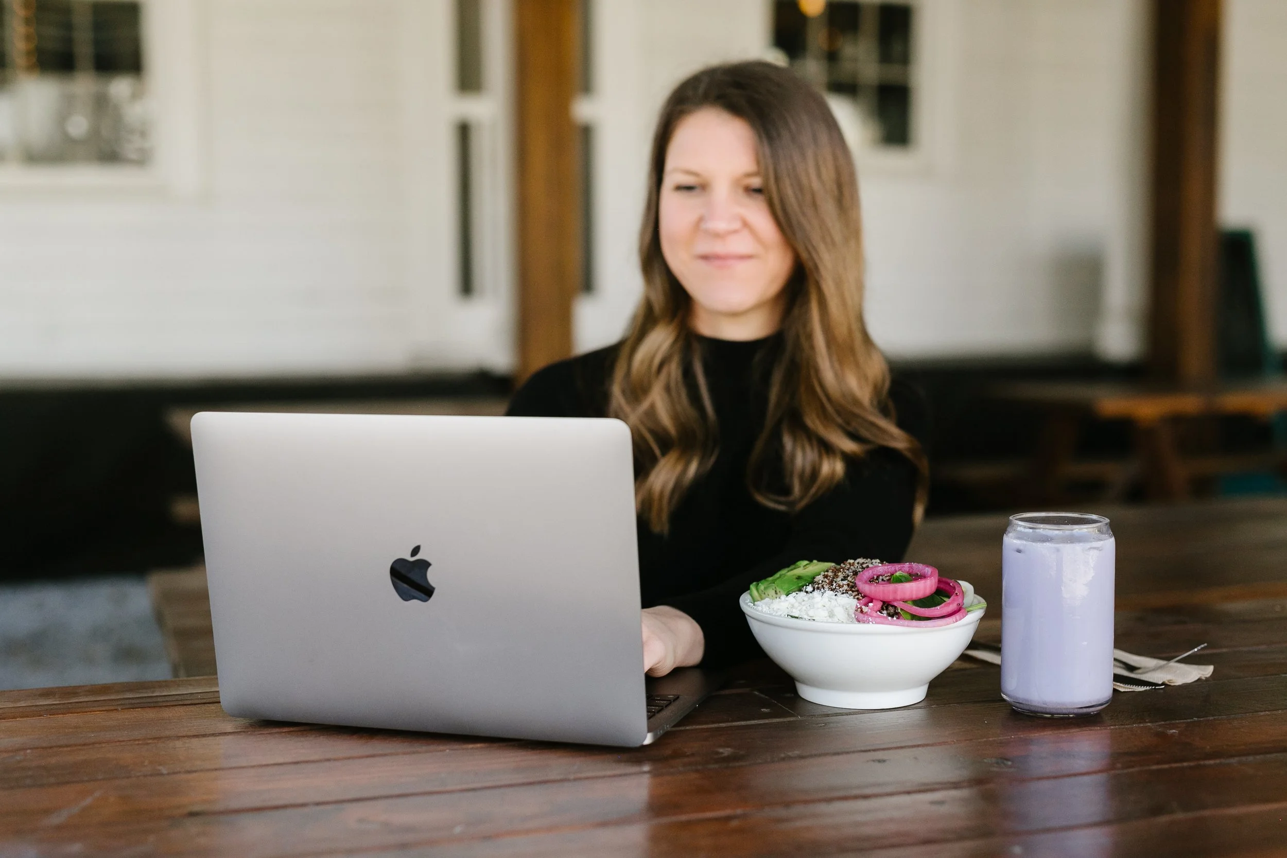 A woman with wavy brown hair working on a laptop at a wooden table, with a bowl of ice cream topped with pink onions, avocados, and sprinkles, and a purple drink next to her in a glass jar.