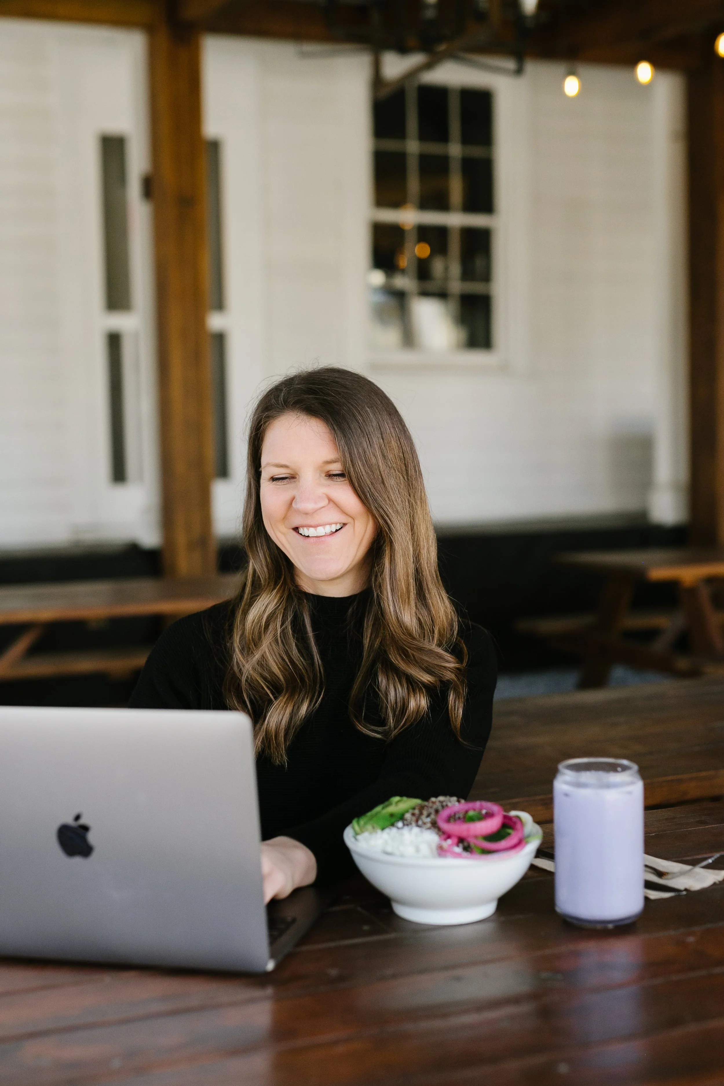 A woman with long brown hair and a black top smiling at her laptop, sitting at a wooden table with a white bowl of salad with onions and vegetables, a glass of water, and a purple candle in a glass holder in front of her, in a cozy indoor space with white walls and a window in the background.