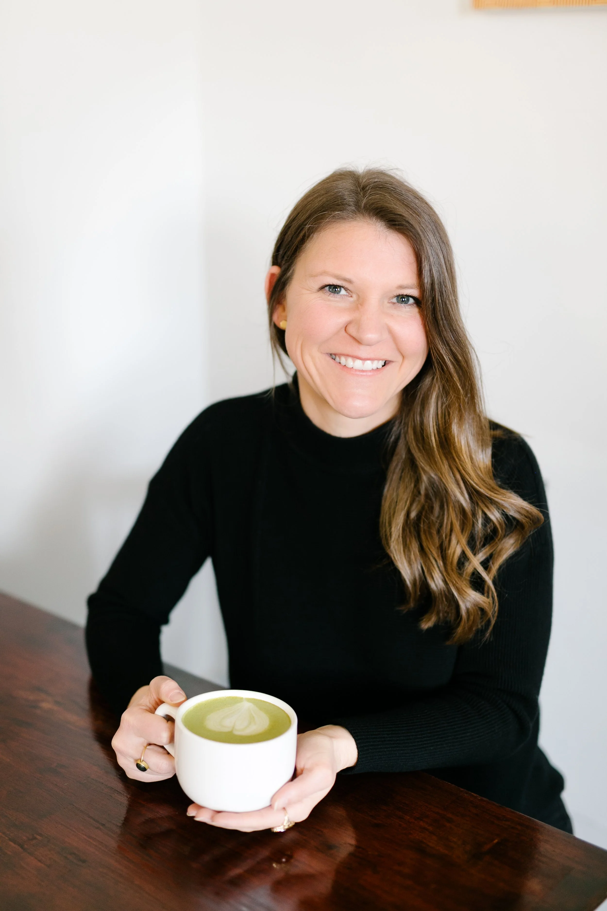 A woman with long brown hair wearing a black sweater, sitting at a wooden table, holding a white mug filled with green matcha latte topped with latte art in the shape of a heart.