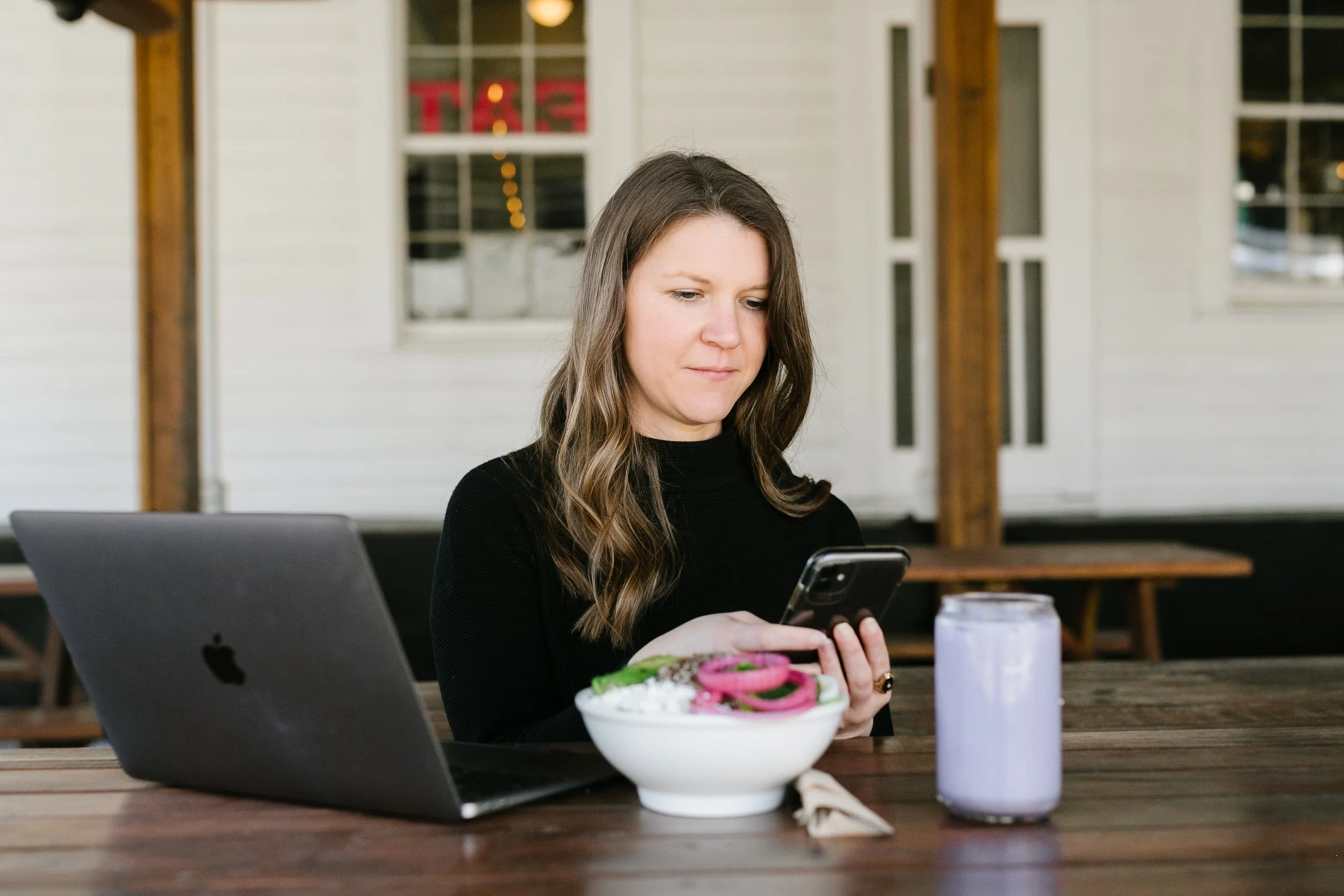 A woman with long brown hair wearing a black top sitting at a wooden table outside, looking at her phone. On the table are a gray open laptop, a bowl of salad with onions and lettuce, a fork, a napkin, a white water bottle, and a purple drink. The background shows a white wooden house with large windows and a porch.