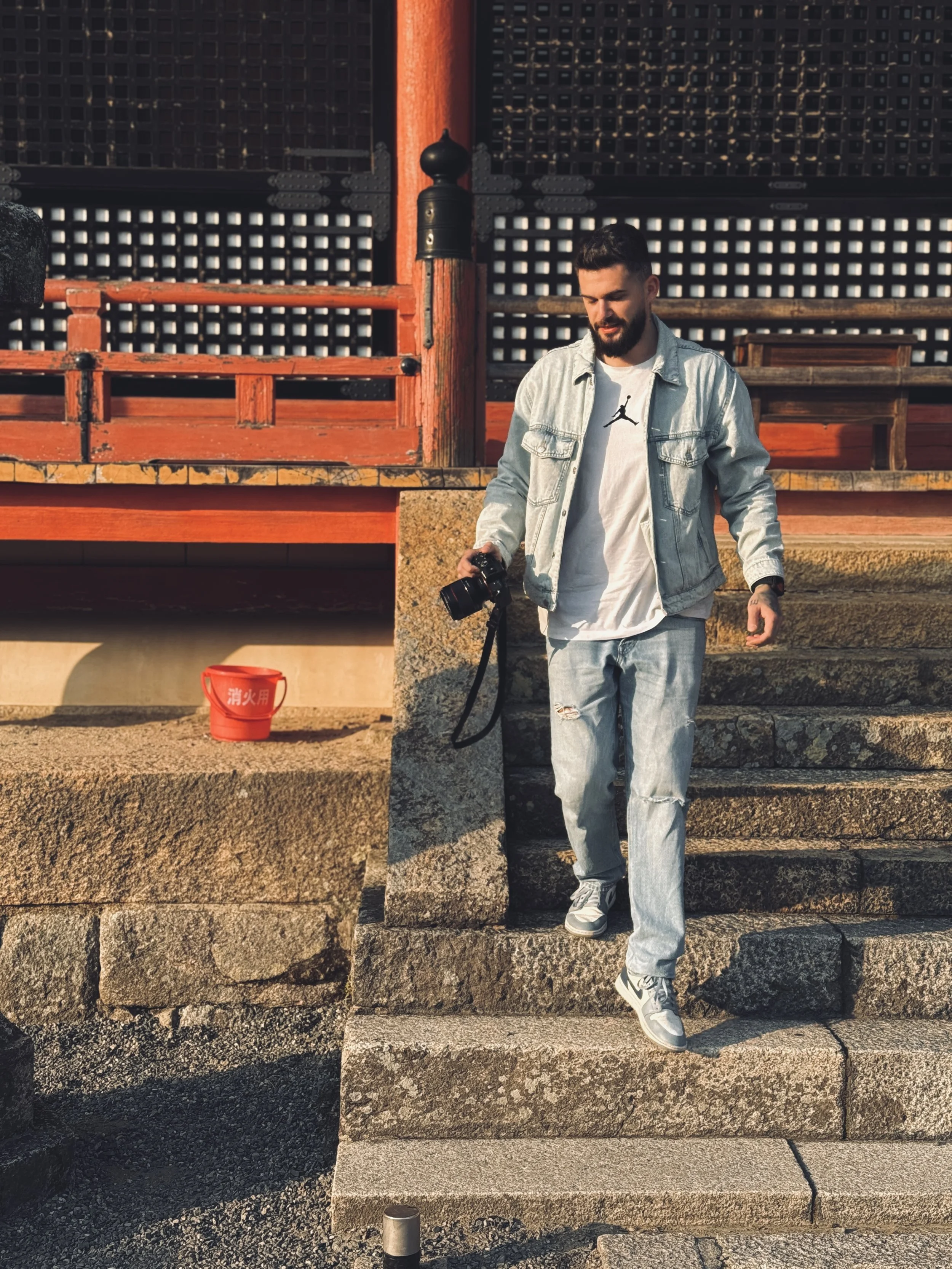 A man with a beard and short dark hair walking down stone steps while holding a camera in his right hand. He is wearing a light denim jacket over a white T-shirt with a black logo, ripped jeans, and sneakers. The background features a traditional wooden building with red and black architectural details and a lattice window. There is a red bucket with white text on the ground behind him.