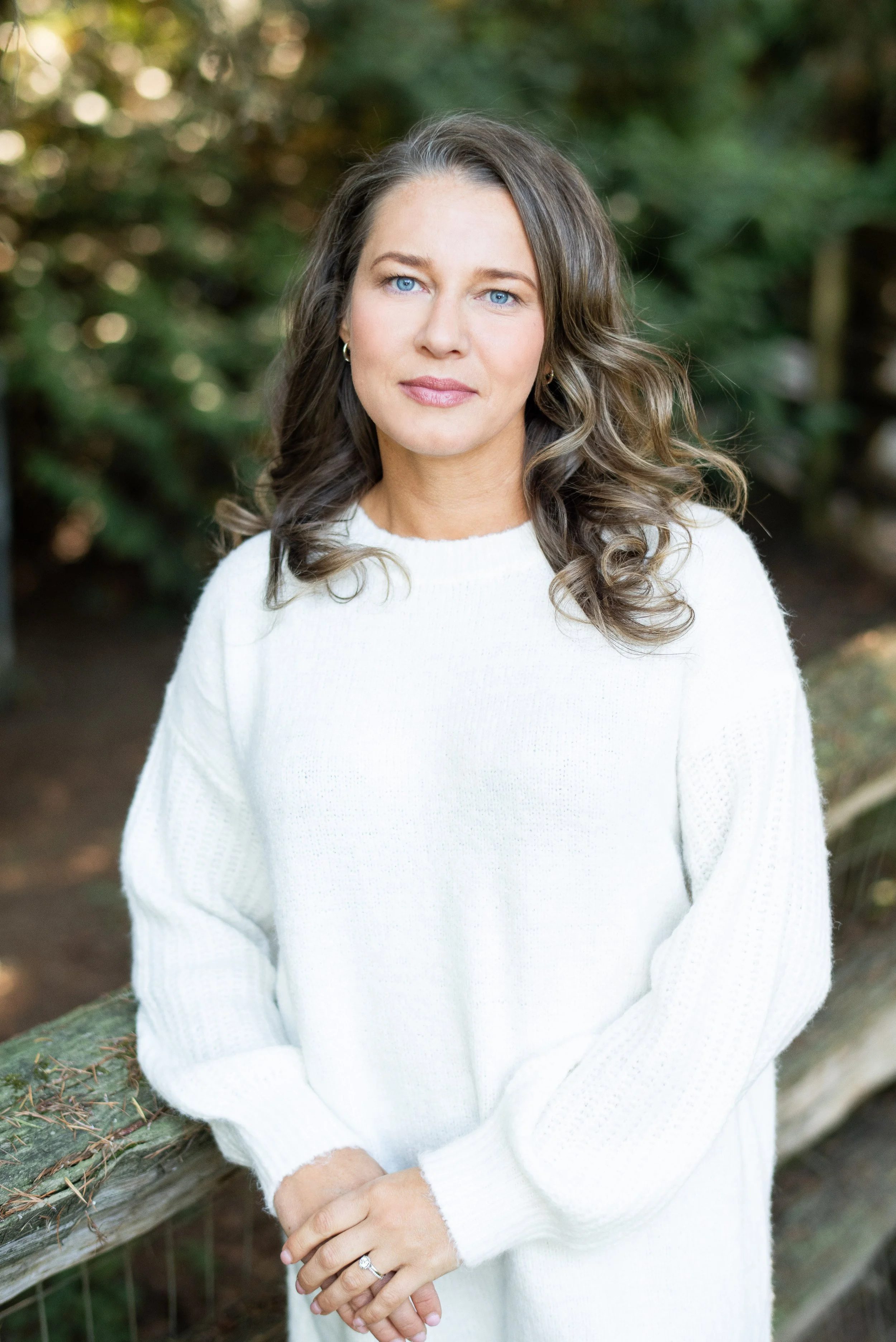 A woman therapist with long, wavy brown hair and blue eyes standing outdoors near a wooden railing with green foliage in the background, wearing a white sweater.