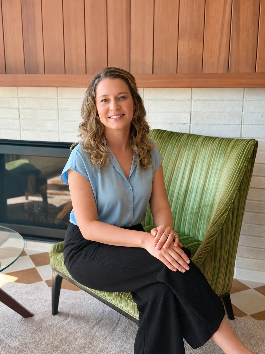 A woman counsellor with curly blonde hair sitting on a green-striped armchair in a therapy room with a wooden and brick fireplace, smiling at the camera.