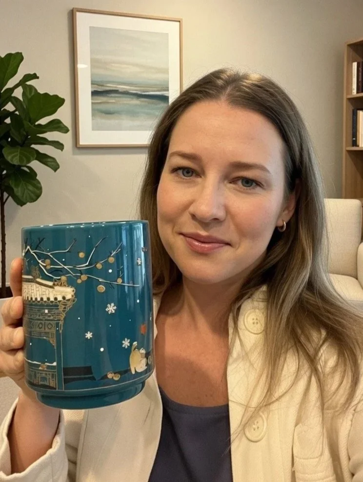 A woman counsellor with long brown hair holding a holiday-themed mug with a winter scene including a bear and building details, standing in a cozy counselling room in Abbotsford with a painting, bookshelf, and plant in the background.