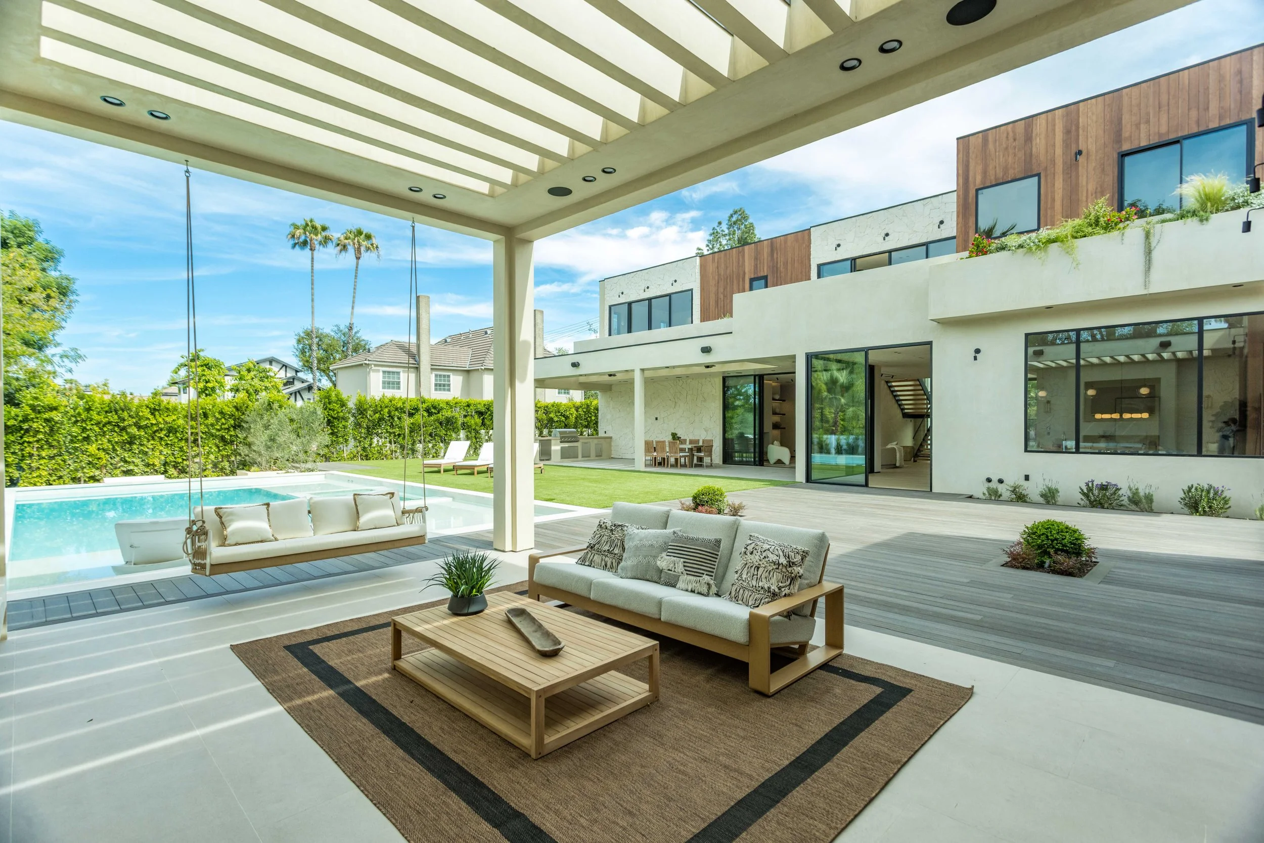 Modern house backyard view with a covered outdoor seating area, pool, outdoor dining and garden, with large windows and a mix of white and wooden exterior walls.