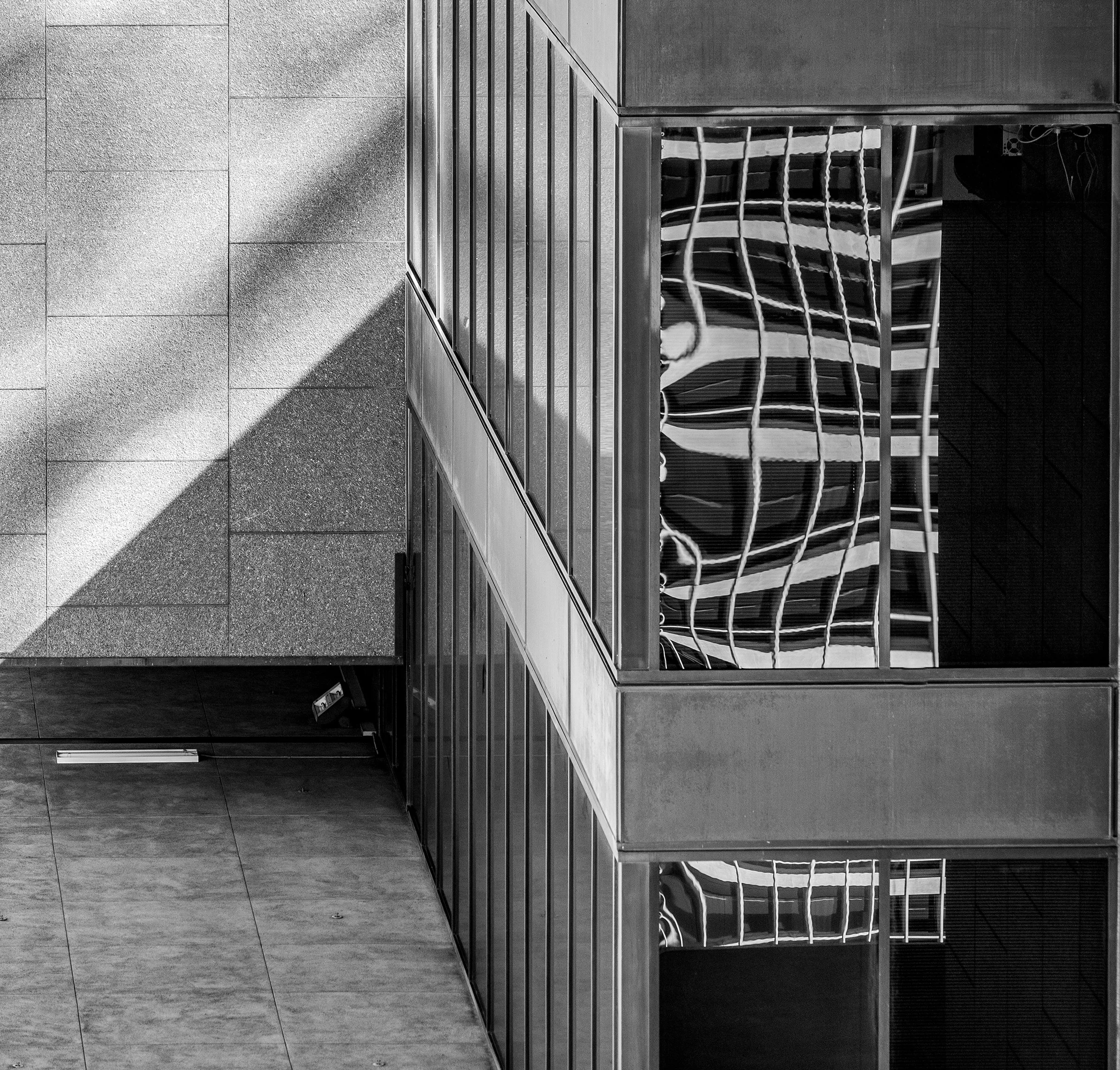 Black and white photo of a modern cityscape reflected in a large glass building, taken outdoors with sunlight casting shadows on the textured ground.