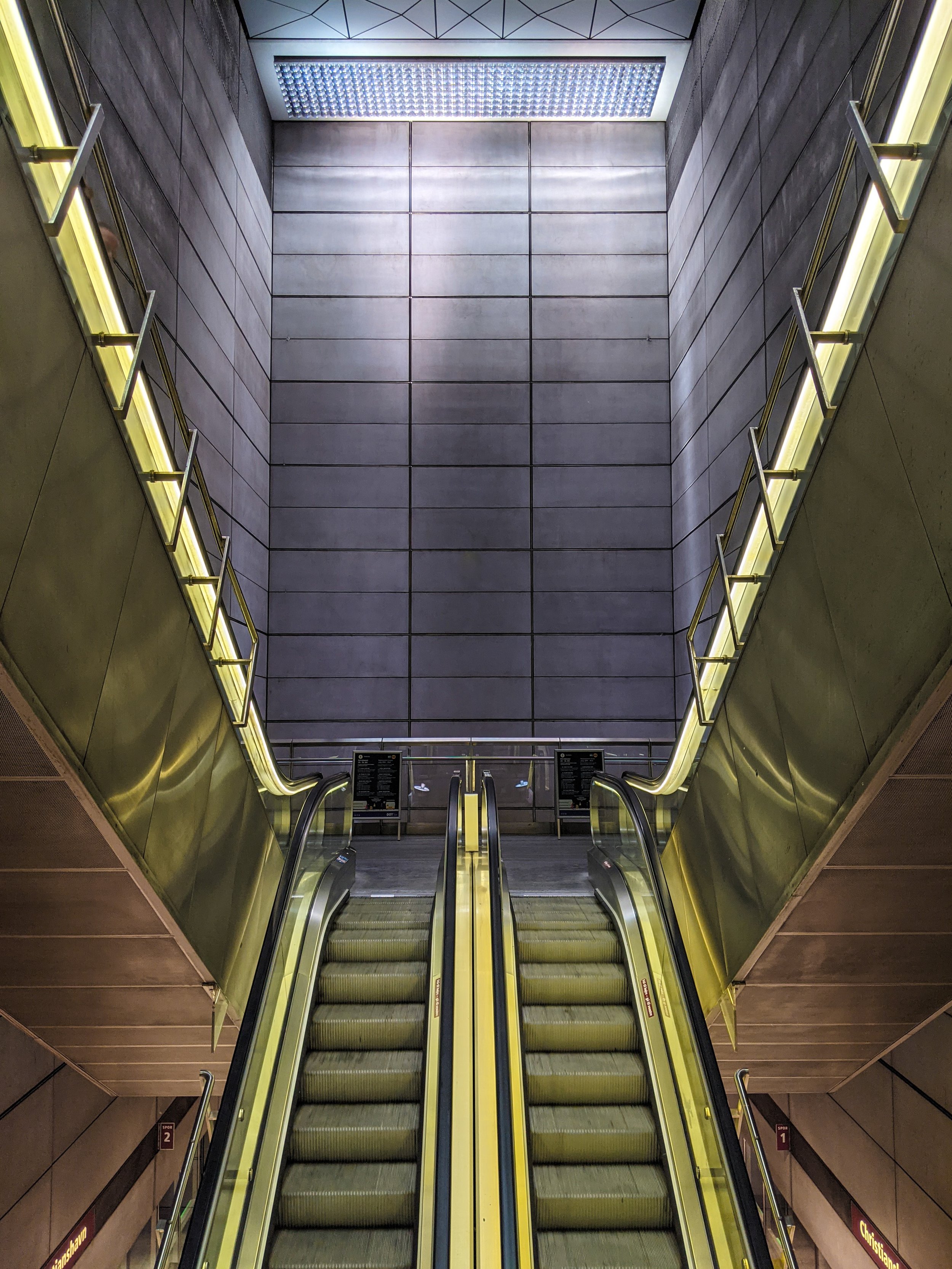 An upward view of escalators leading to an underground station with metal panel walls and a large illuminated ceiling grille.
