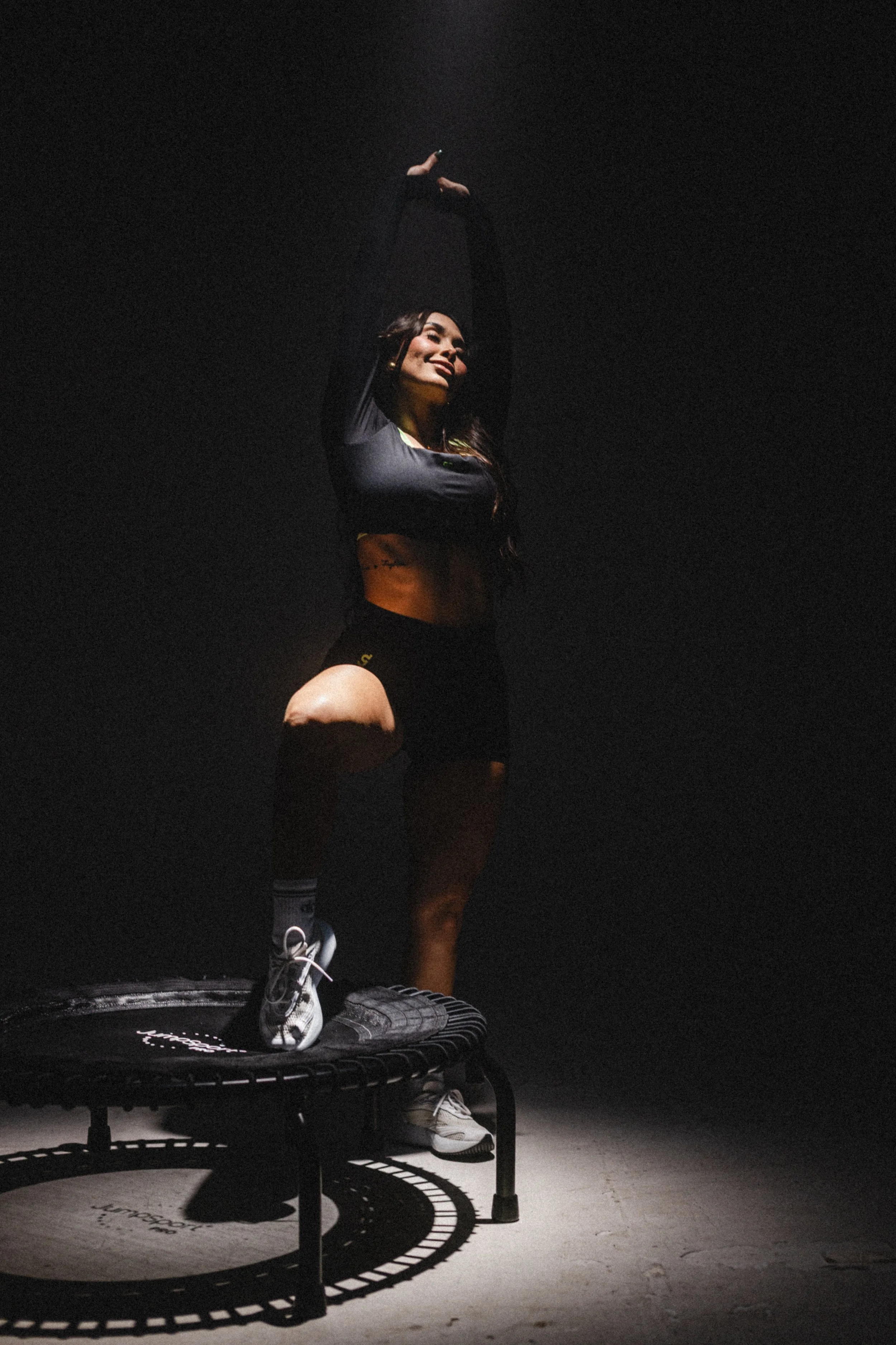 A woman doing a sports stretch or yoga pose on a trampoline in a dark room, with dramatic lighting highlighting her smiling face, athletic black clothing, and sports shoes.