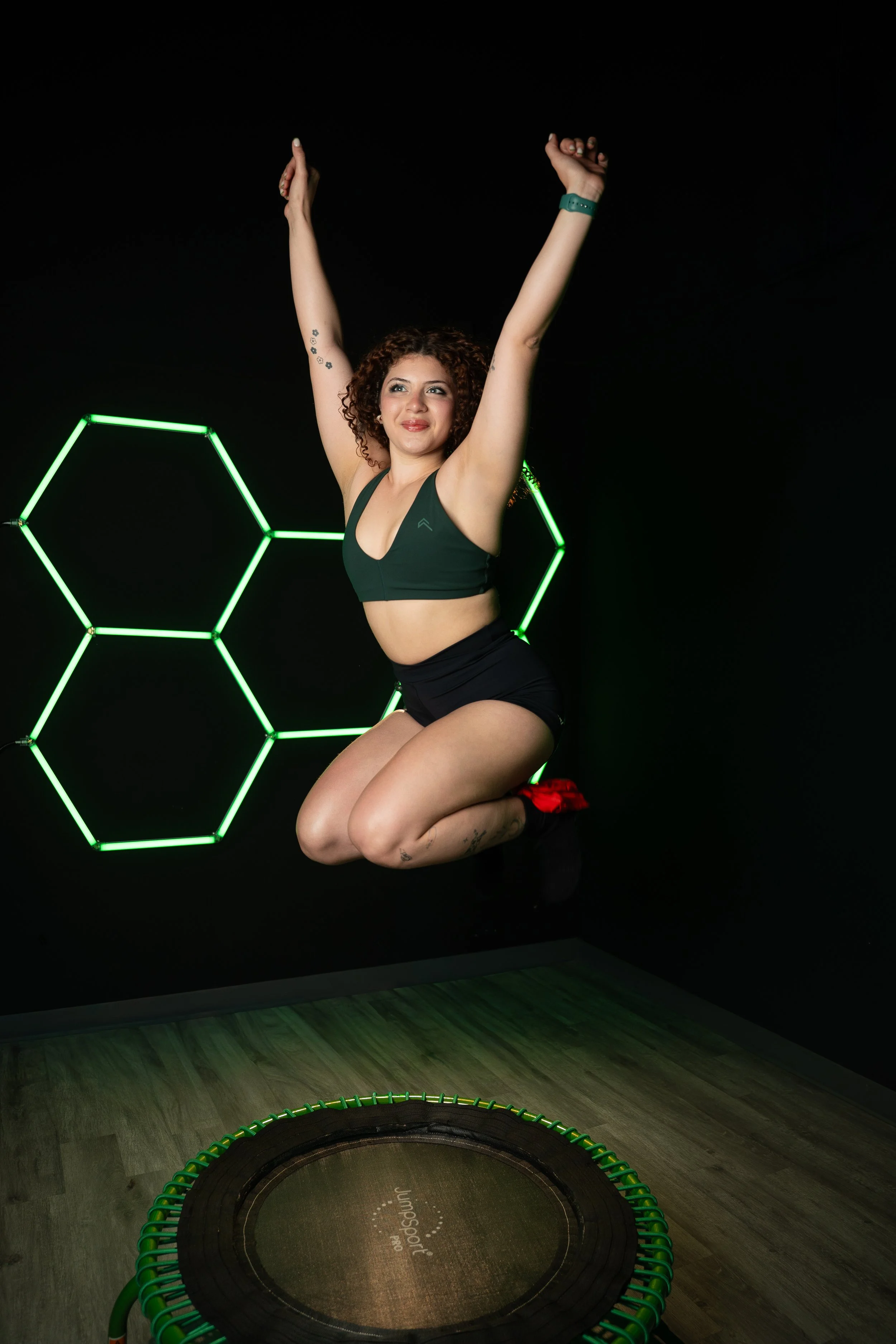 A woman with curly hair, wearing a dark sports bra and shorts, is mid-jump on a trampoline with a black and green frame, inside a room with black walls and neon green hexagon shapes on the wall.