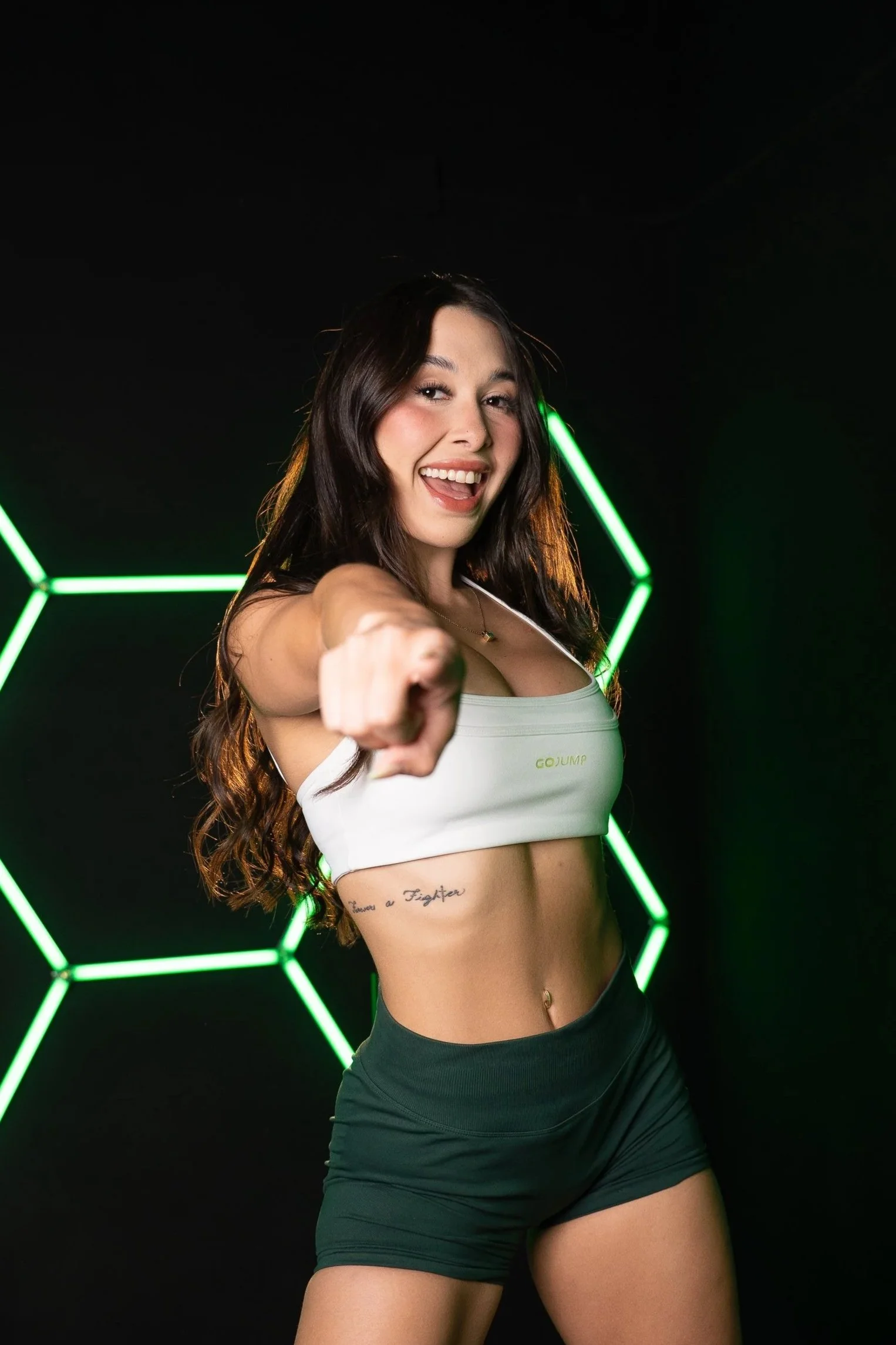 A young woman with long dark hair smiling and pointing towards the camera in front of black background with green neon hexagon shapes.