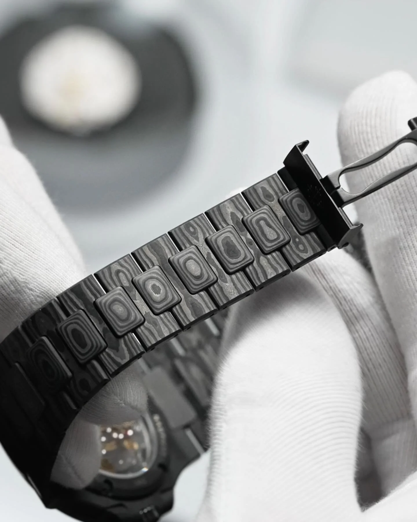 Close-up of a luxury wristwatch with a patterned metal band, held by a gloved hand against a blurred background.