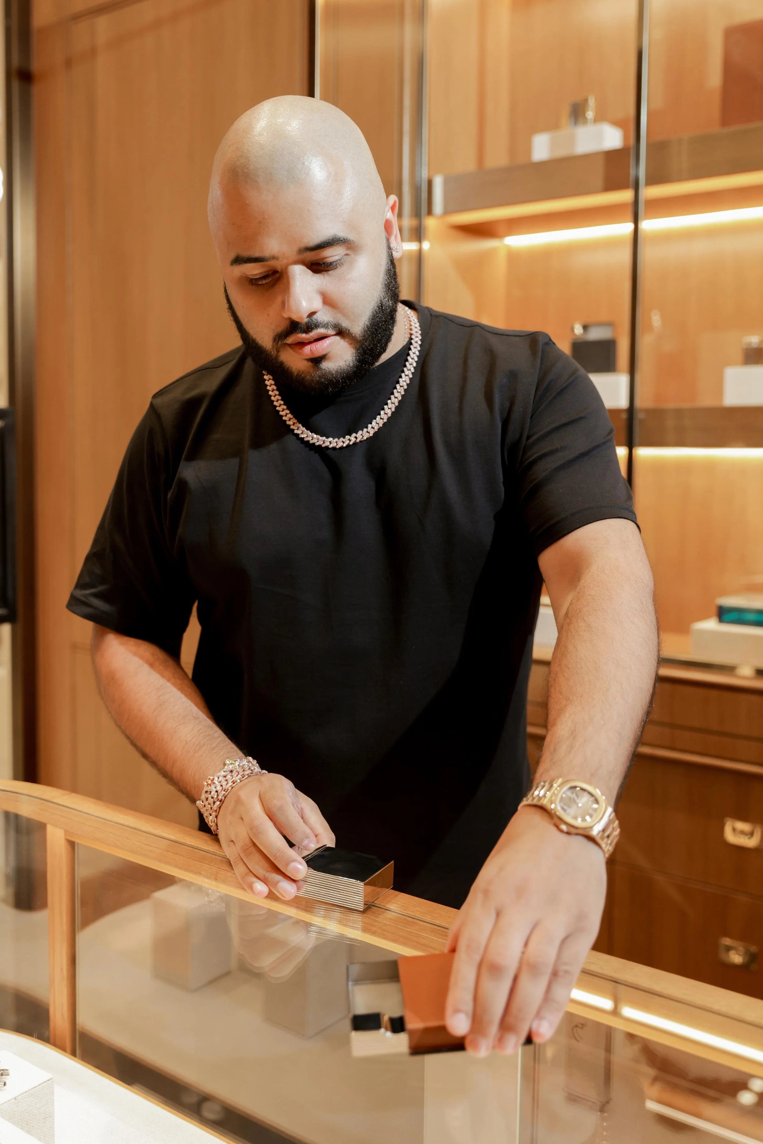 A bald man with a beard, wearing a black shirt, gold watch, and jewelry, is looking down and handling jewelry or items in a jewelry store with wooden shelves in the background.