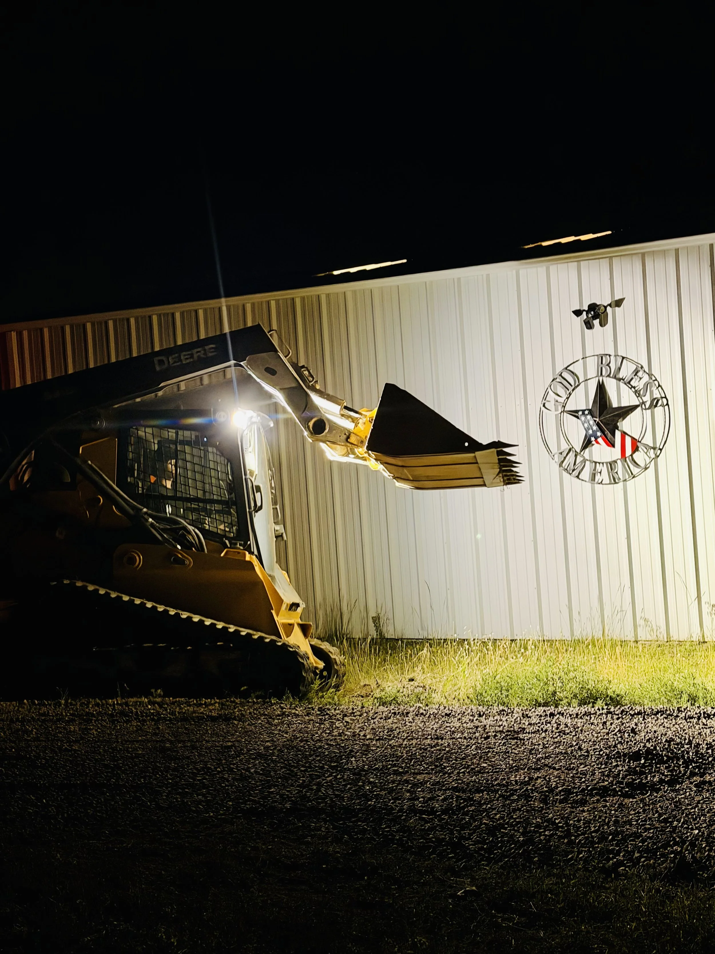 Nighttime scene showing a yellow John Deere skid steer loader parked on gravel in front of a white metal building. The building has a Large round clock with the words 'Good Beer' and 'Merica' along with a star. There is a beige patio umbrella and a security camera mounted on the wall.