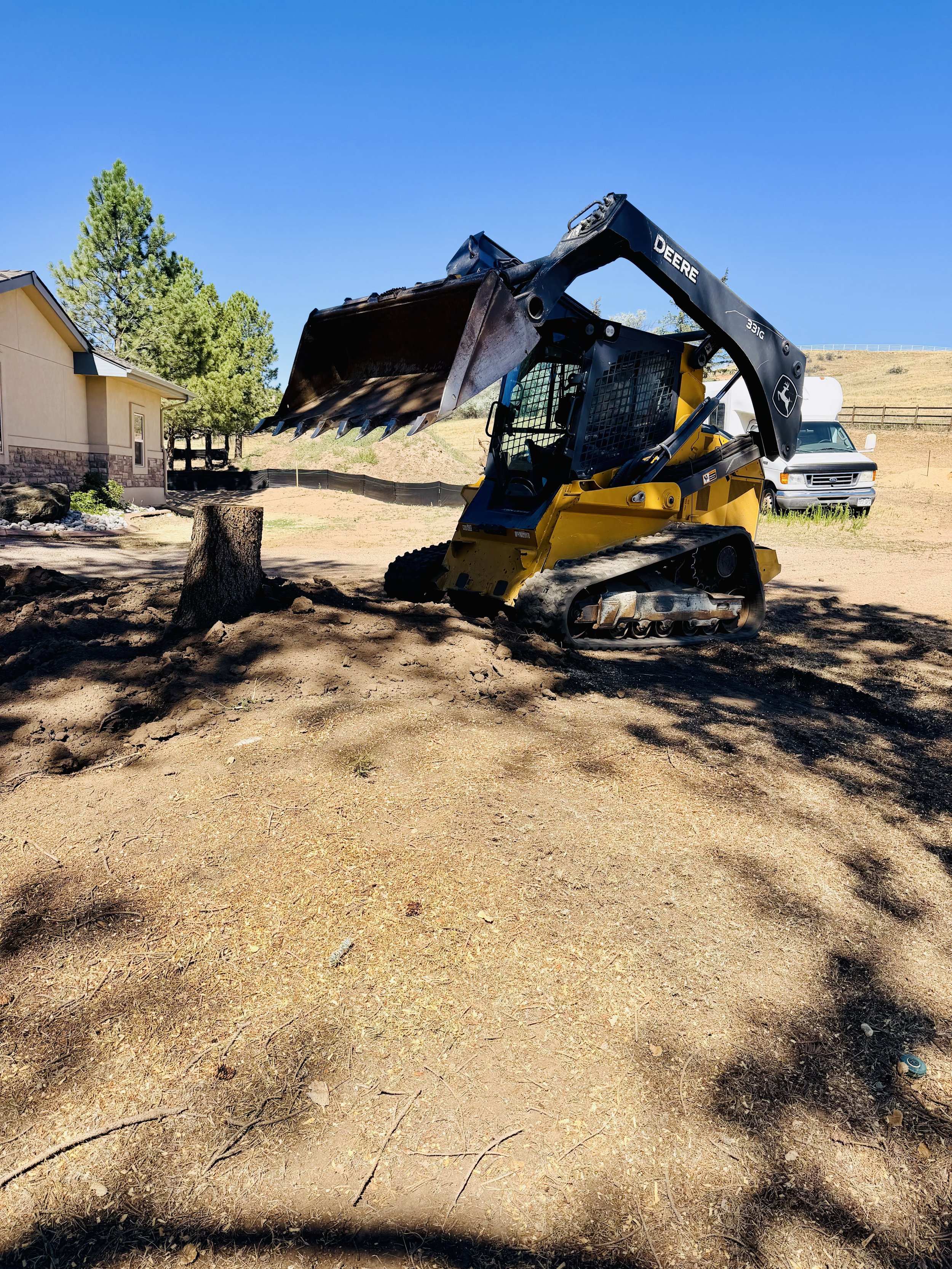 A construction site with a small tracked excavator on dirt, near a tree and a house, under clear blue sky with some distant hills and a van in the background.