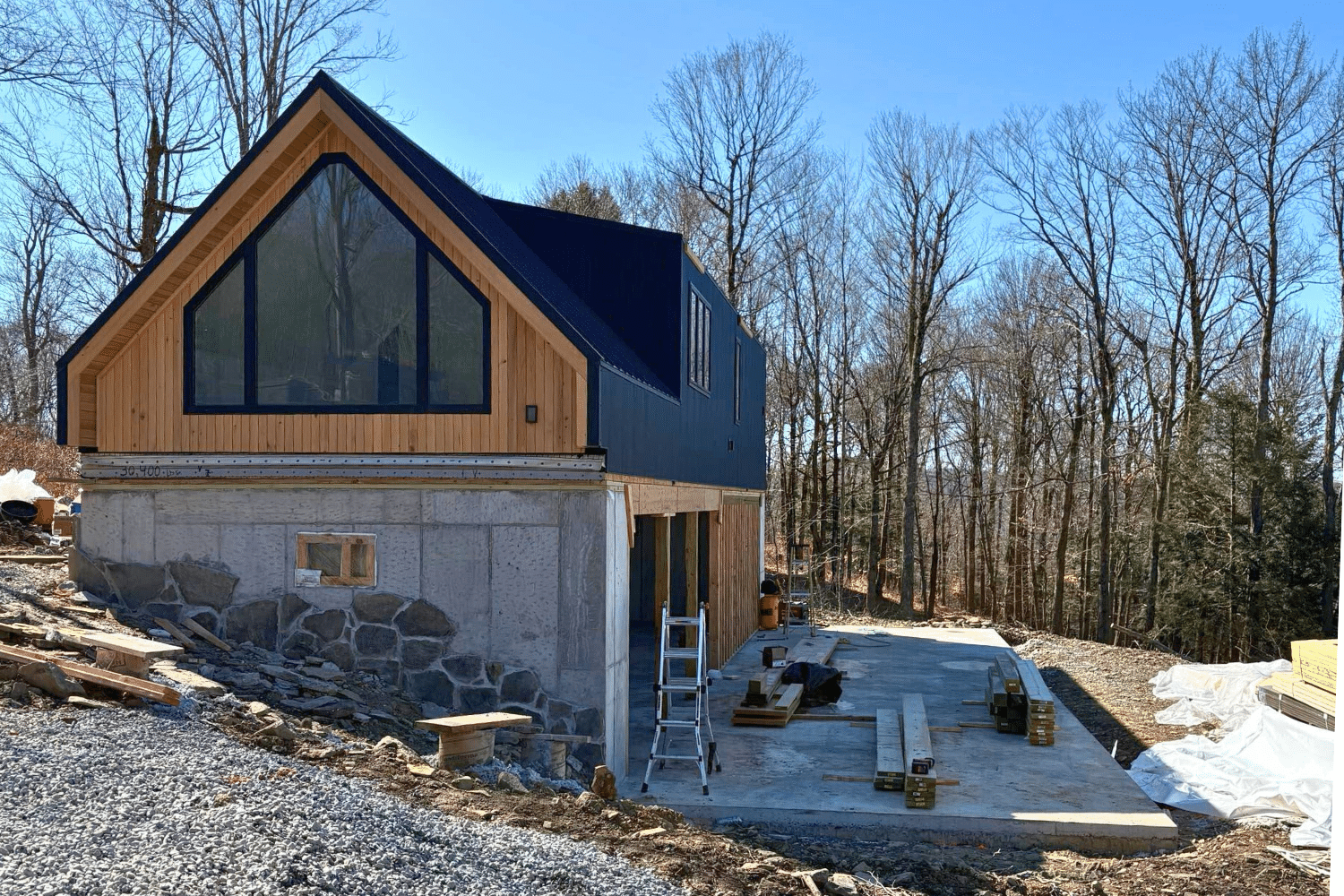 A two-story house under construction with a wooden upper level and stone foundation, large front window, surrounded by construction tools and materials, with leafless trees in the background on a clear, sunny day.