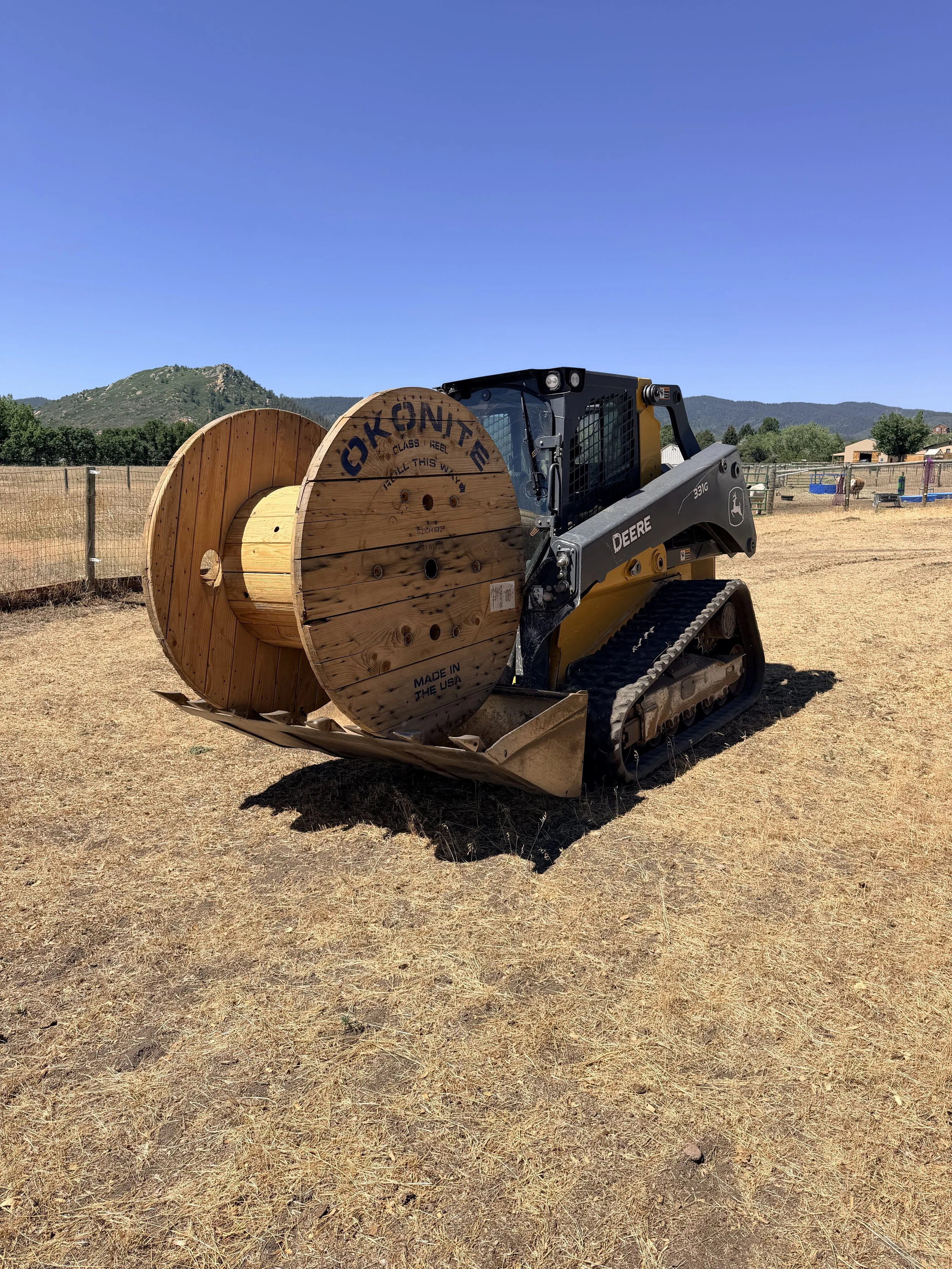 A small John Deere skid steer loader with black and yellow colors lifting a large wooden spool with the word "OKONTE" painted on it in black. The background features a dry, grassy field, a wooden fence, trees, small structures, and distant green mountains under a clear blue sky.