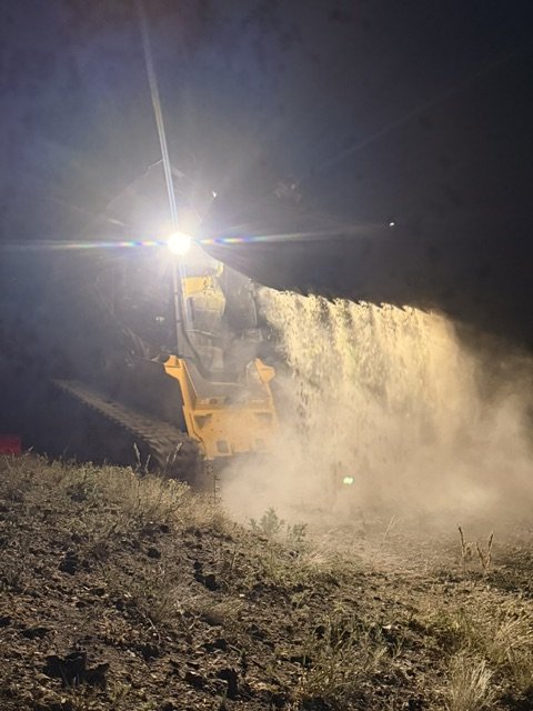 A construction excavator operating at night, pouring dirt or soil, with bright headlights illuminating the scene.