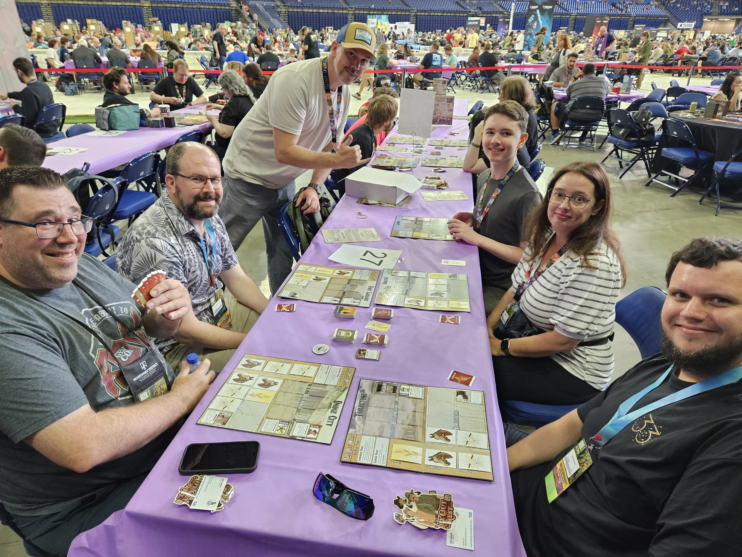 Group of people playing a tabletop board game at a convention or event, with many tables, chairs, and other participants visible in the background. The players are seated around a long table covered with a purple cloth, engaging with the game components, cards, and tokens.