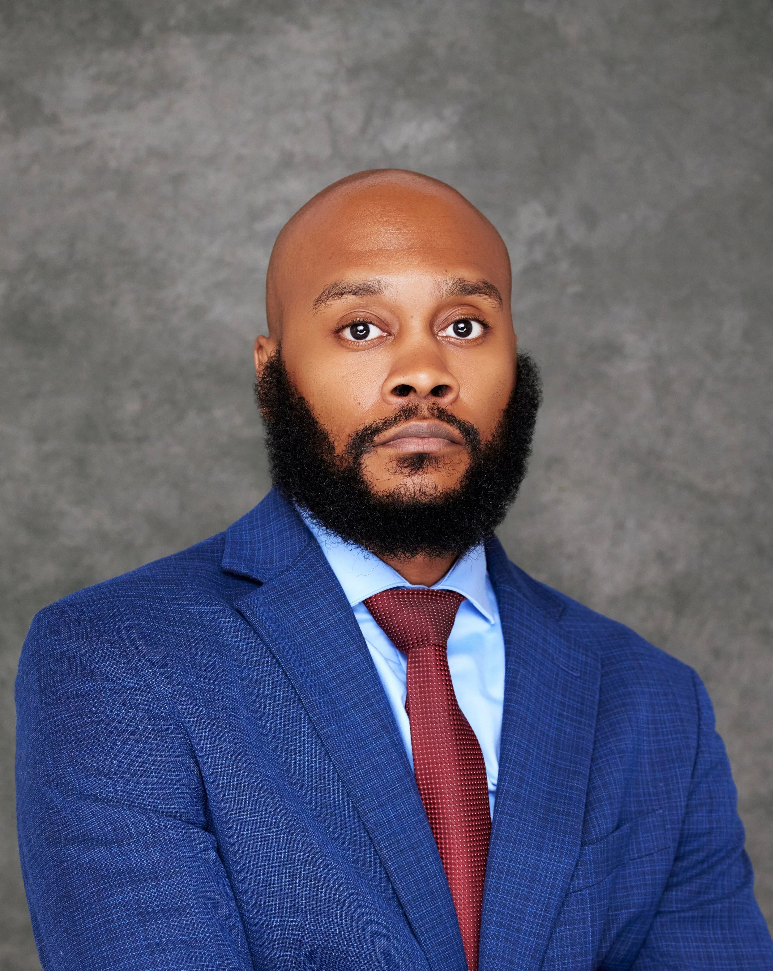A close-up portrait of a man with a dark skin tone, bald head, and full beard, wearing a blue checked suit, a light blue shirt, and a red tie, against a gray textured background.