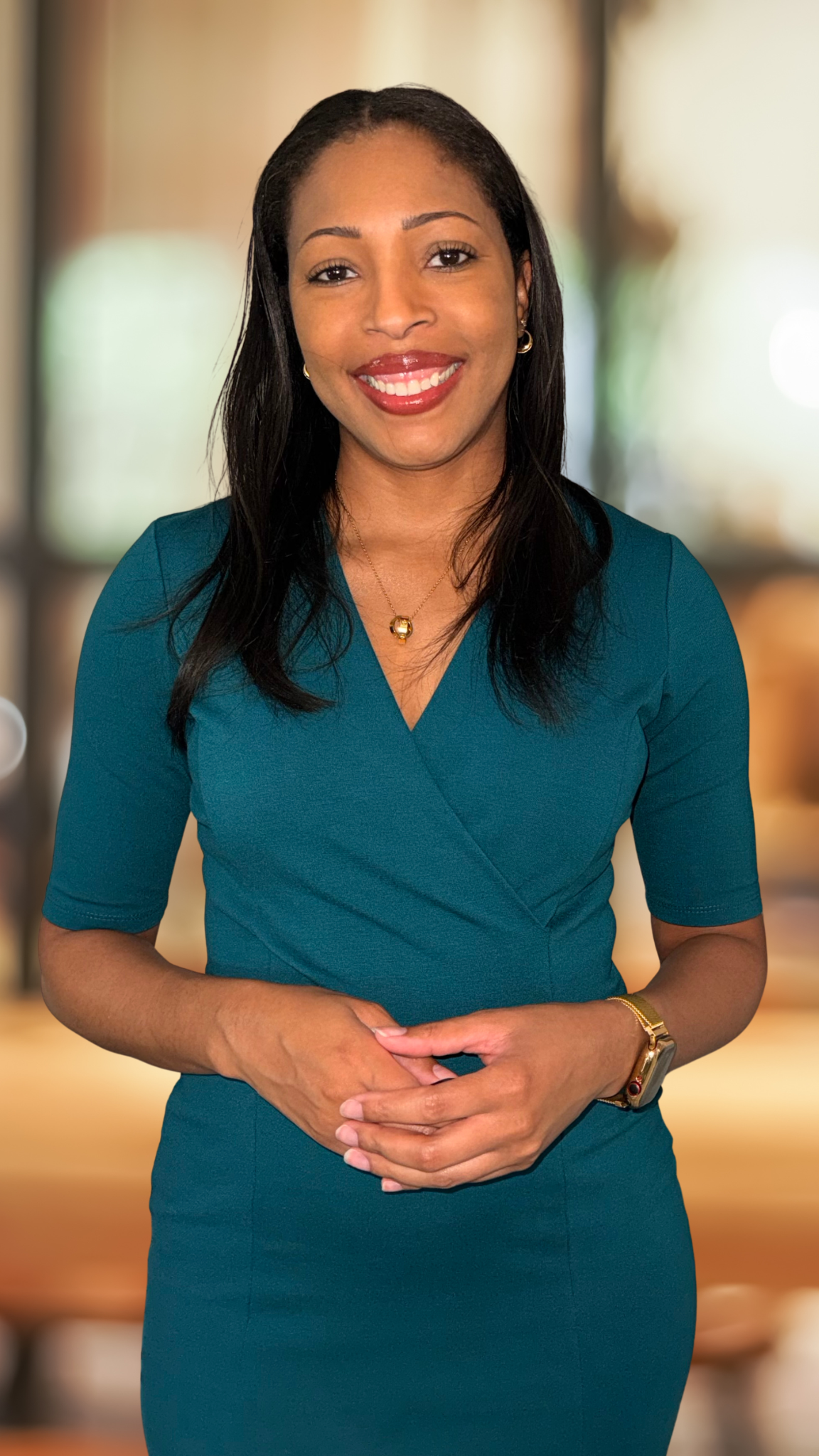 A smiling woman with medium skin tone and black hair, wearing a teal dress, gold jewelry, and a watch, standing indoors with a blurred background.