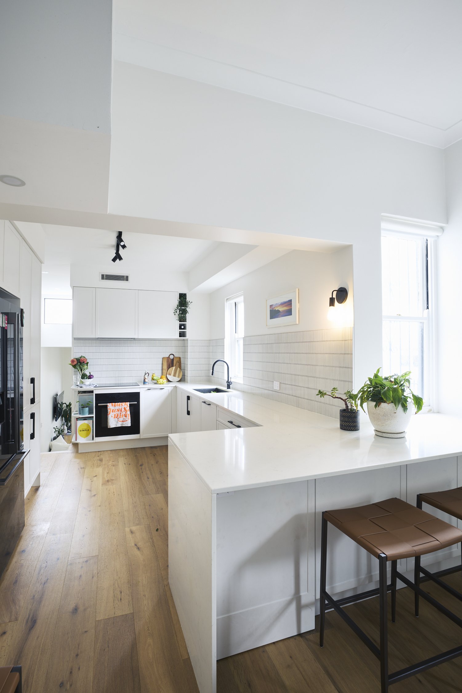 Bright modern kitchen in a Victorian townhouse with white cabinets, a marble island, and wooden floors. Potted plants and a wall art provide decor, with ample natural light from large windows.