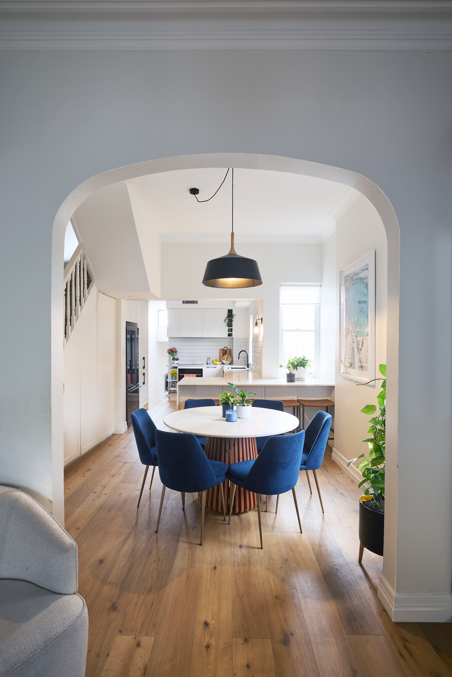 Open-concept dining area with a round white table surrounded by six dark blue upholstered chairs, wooden flooring, a black pendant light, and a view into the kitchen with white cabinets, a window, and a plant.
