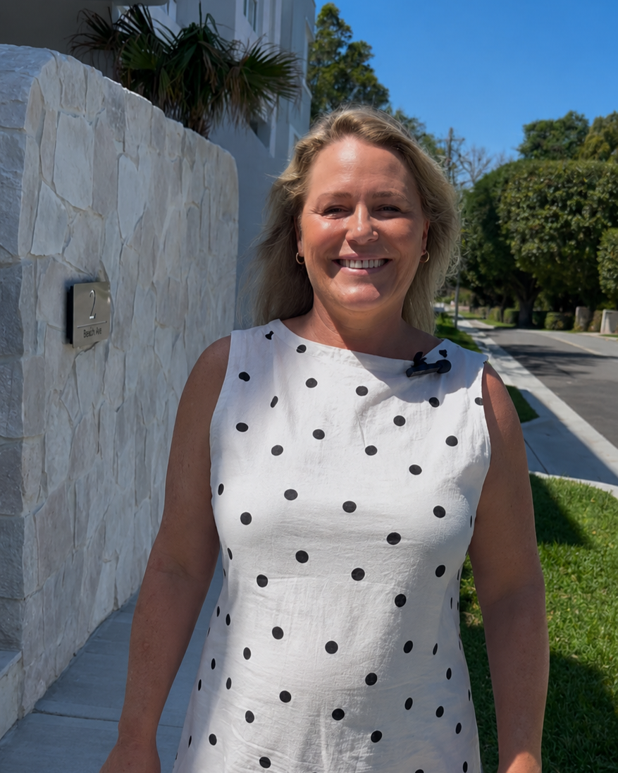 Interior designer smiling in a white dress with black polka dots standing outside a newly renovated house on a sunny day, near a stone wall and greenery.