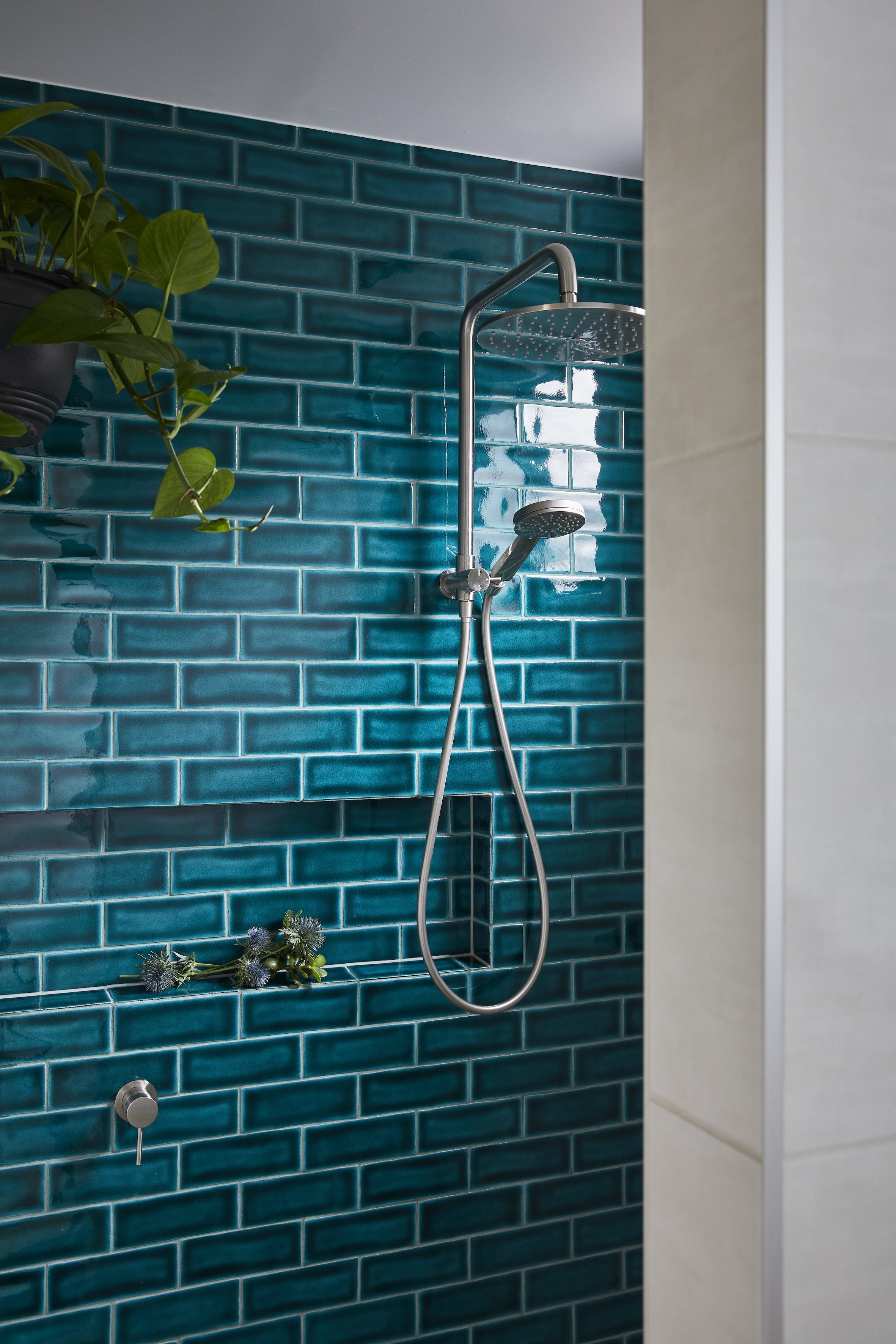 A shower with emerald subway tiles, a rainfall showerhead, and a handheld showerhead, with a green plant and flowers on the shelf.